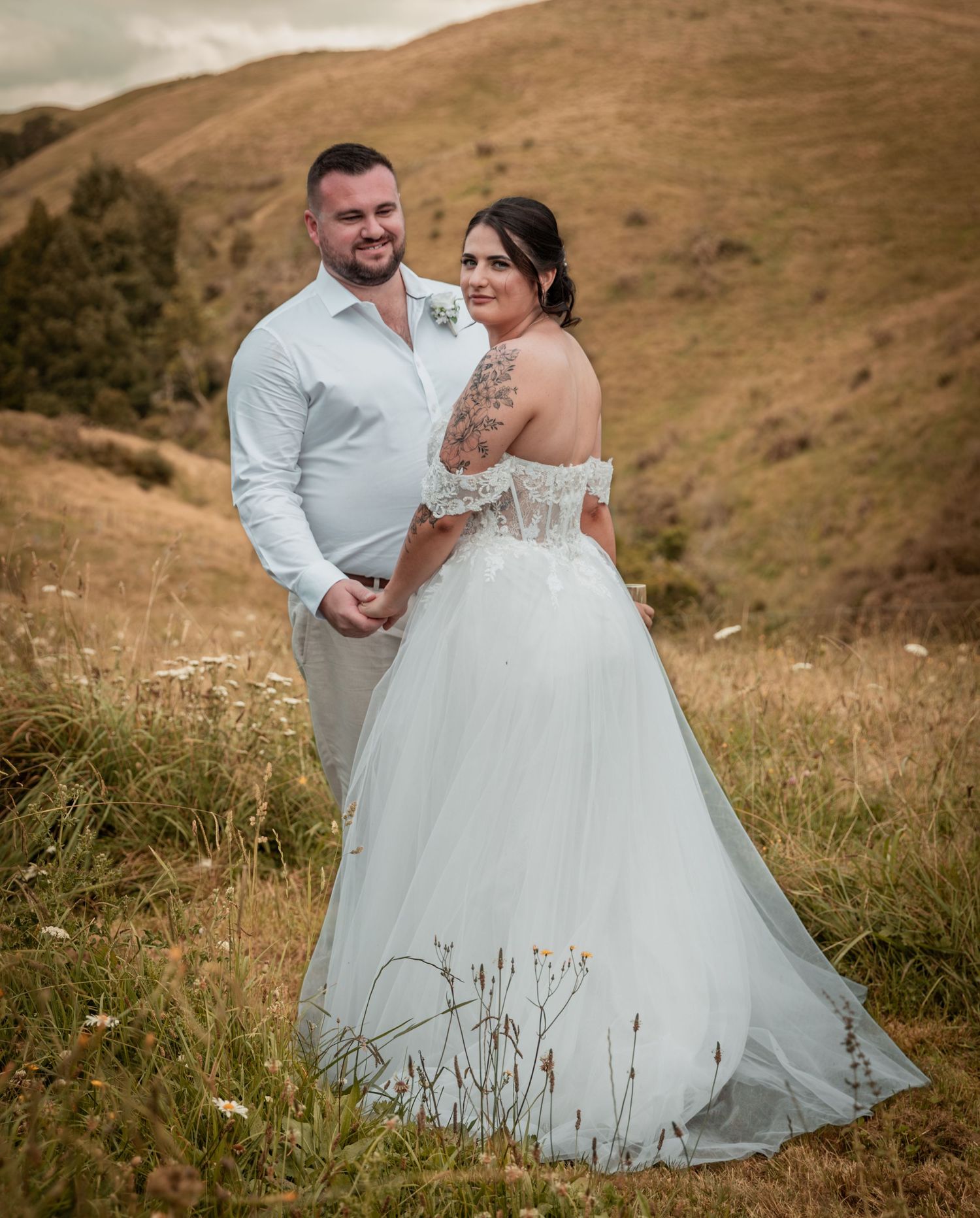 Bride in an off-shoulder lace ballgown and groom in a white shirt posing on a scenic green hillside.