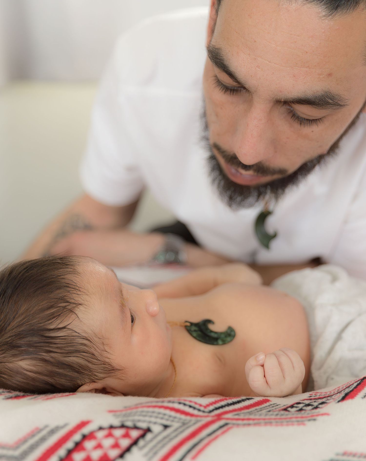 Tattooed father gazing lovingly at sleeping newborn baby with a greenstone Maori pendant placed on chest.