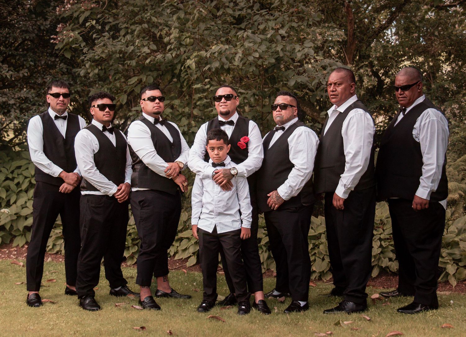 Groomsmen in black vests and bow ties posing with young ring bearer on green lawn.