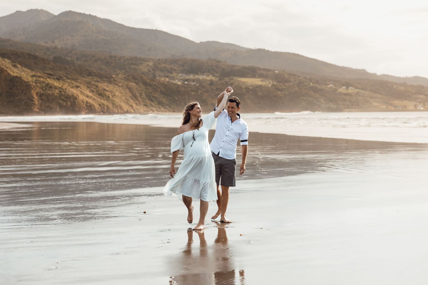 Couple dances barefoot on reflective wet beach sand with scenic mountain backdrop at golden hour.