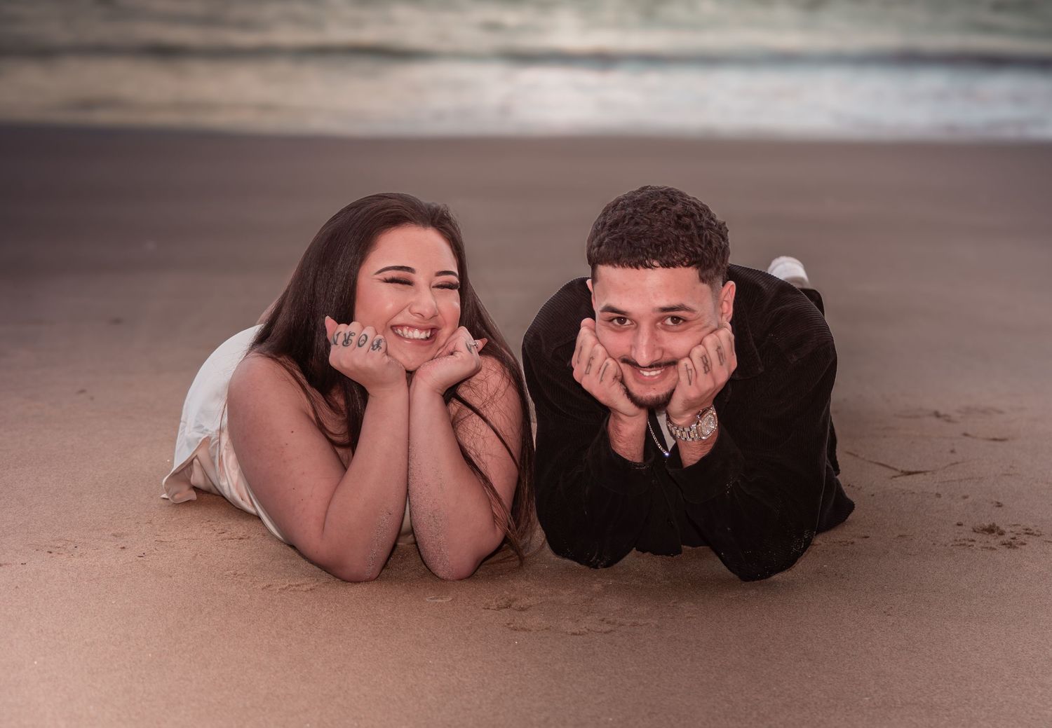 Happy couple lying on sandy beach smiling in casual black and white engagement photo.