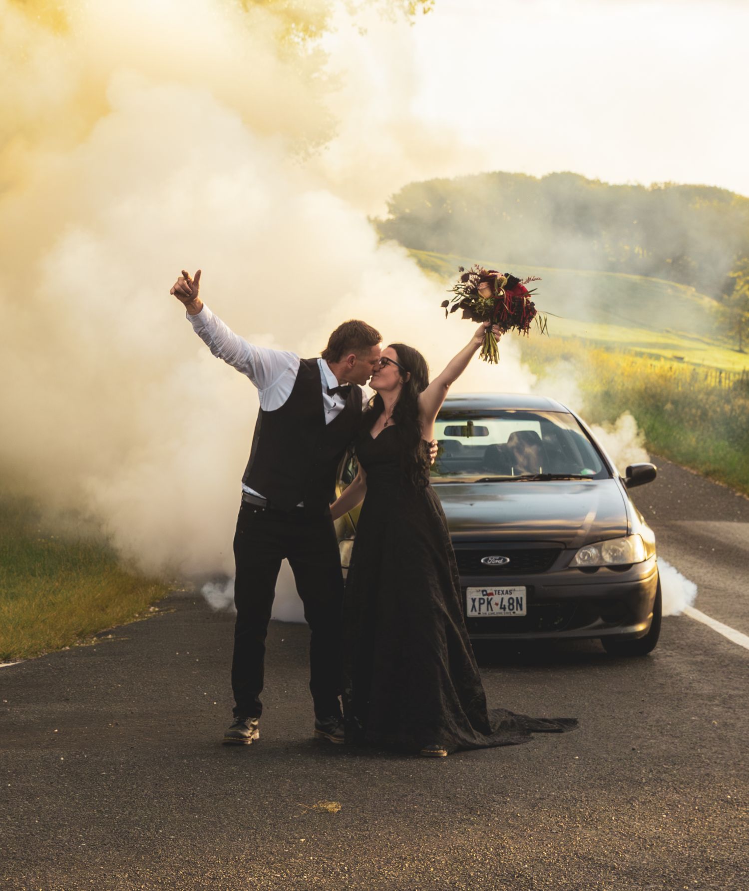 Couple kisses in front of car with colorful smoke bombs on road, bride in black gown holds bouquet.