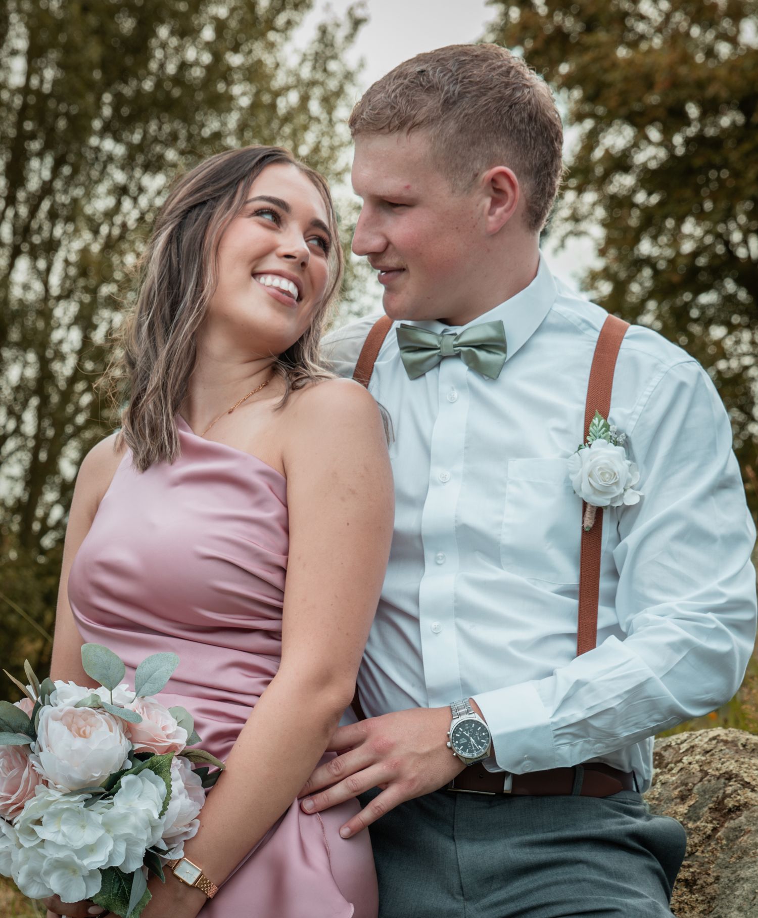 Smiling couple in wedding attire, pink dress and sage bow tie with suspenders, pose outdoors.