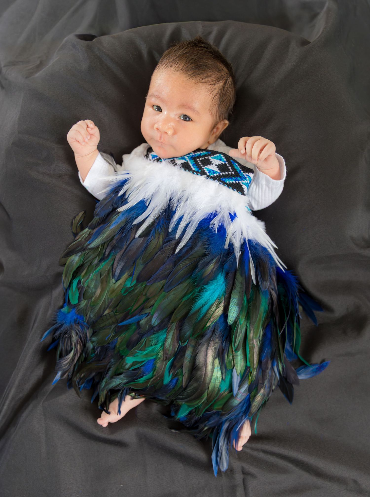 Newborn baby lying on dark fabric wearing a traditional Maori feather cloak with blue and green plumage.