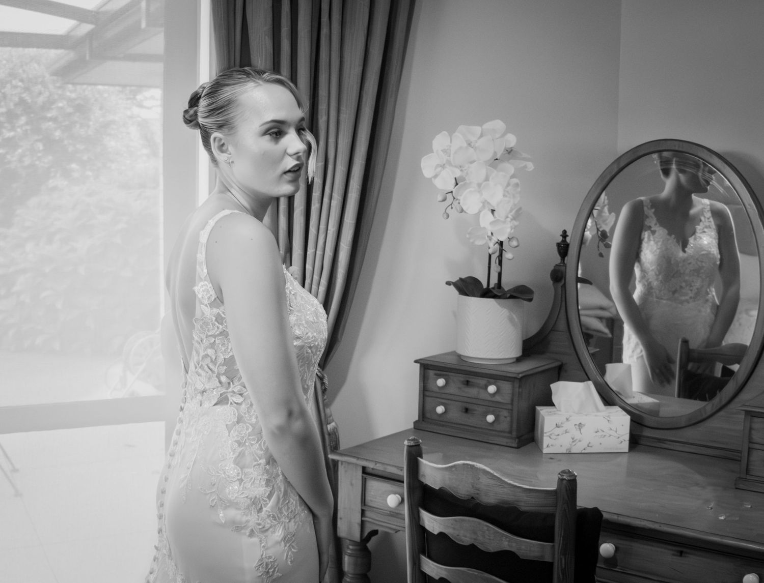 Bride in lace wedding gown stands by window with mirror reflection in black and white photo.