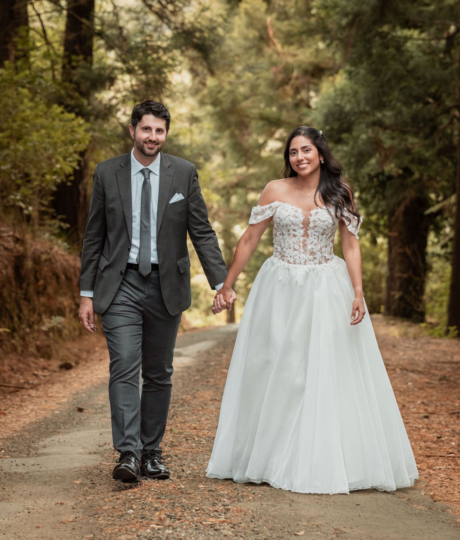 Bride in off-shoulder floral gown and groom in grey suit walking hand in hand along forest path.