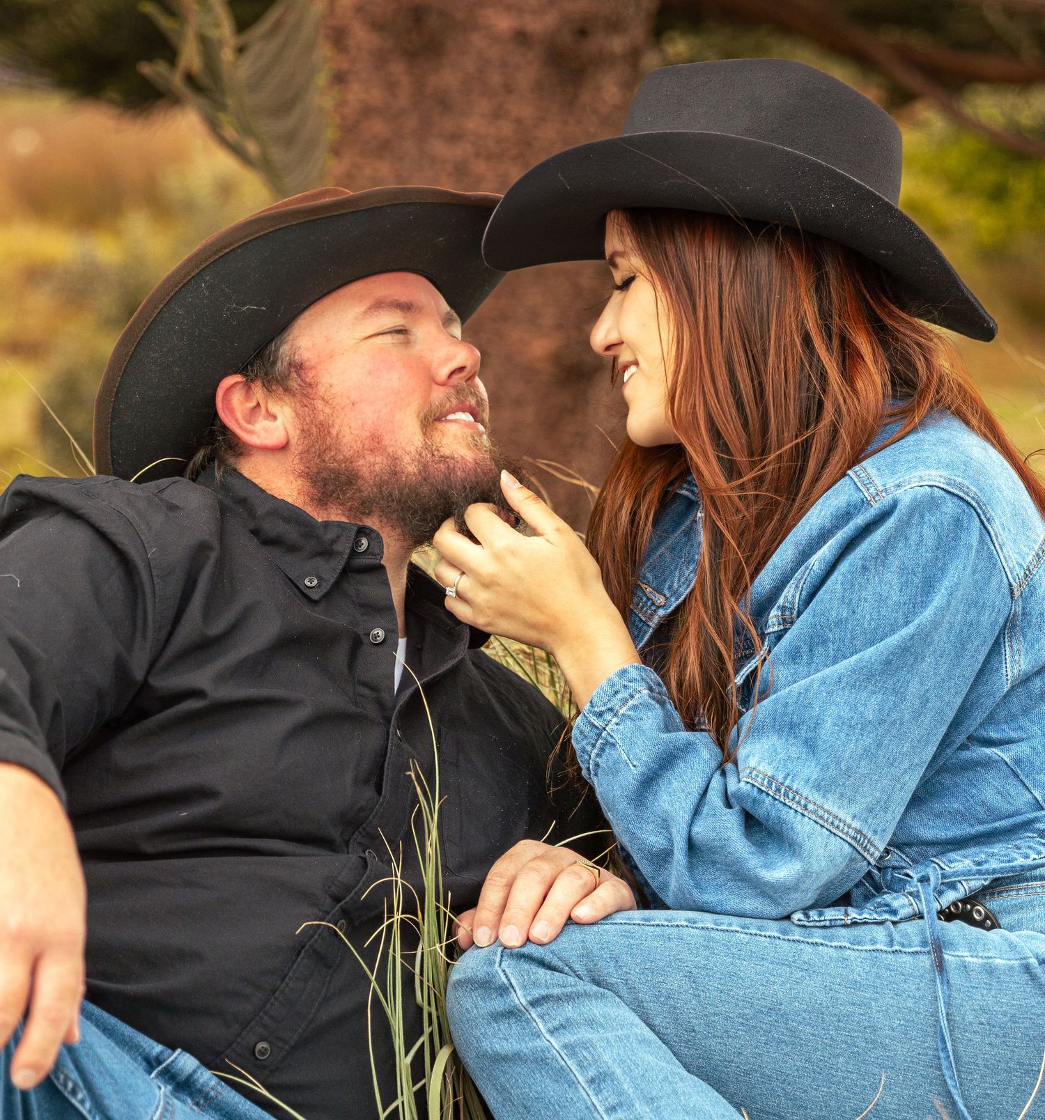 Romantic couple in matching black cowboy hats sitting outdoors in a field, smiling and gazing at each other.