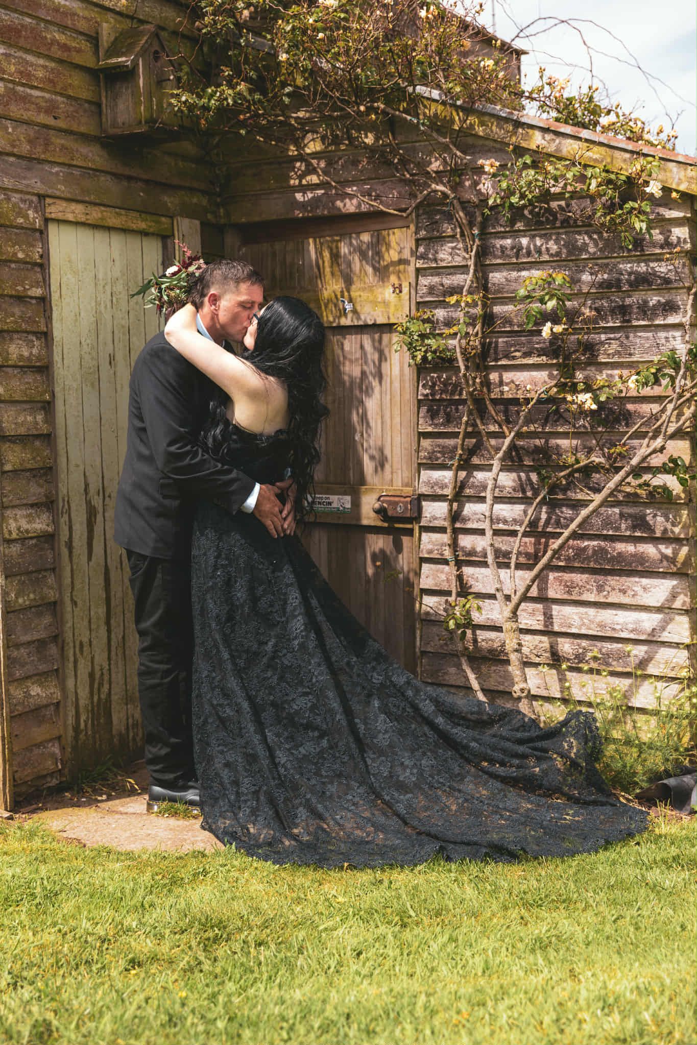 Bride in black lace gown and groom share romantic kiss outside rustic wooden shed with climbing vines.