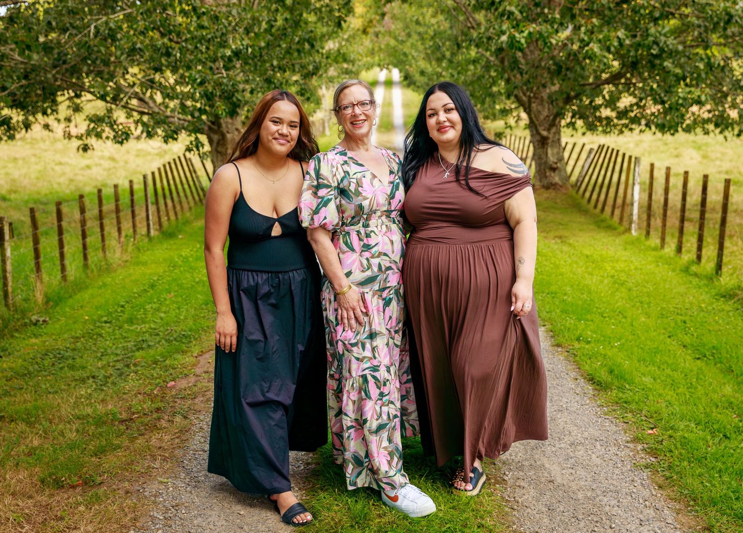 Three women smile together on a tree-lined garden path in a outdoor portrait photo.