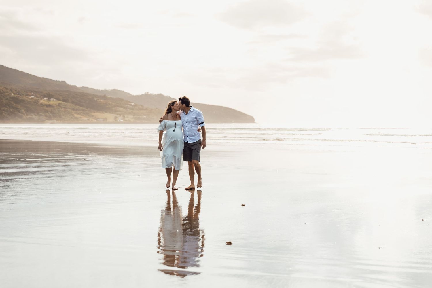 A romantic couple walks barefoot on a reflective beach at sunset, sharing a tender kiss near the ocean.
