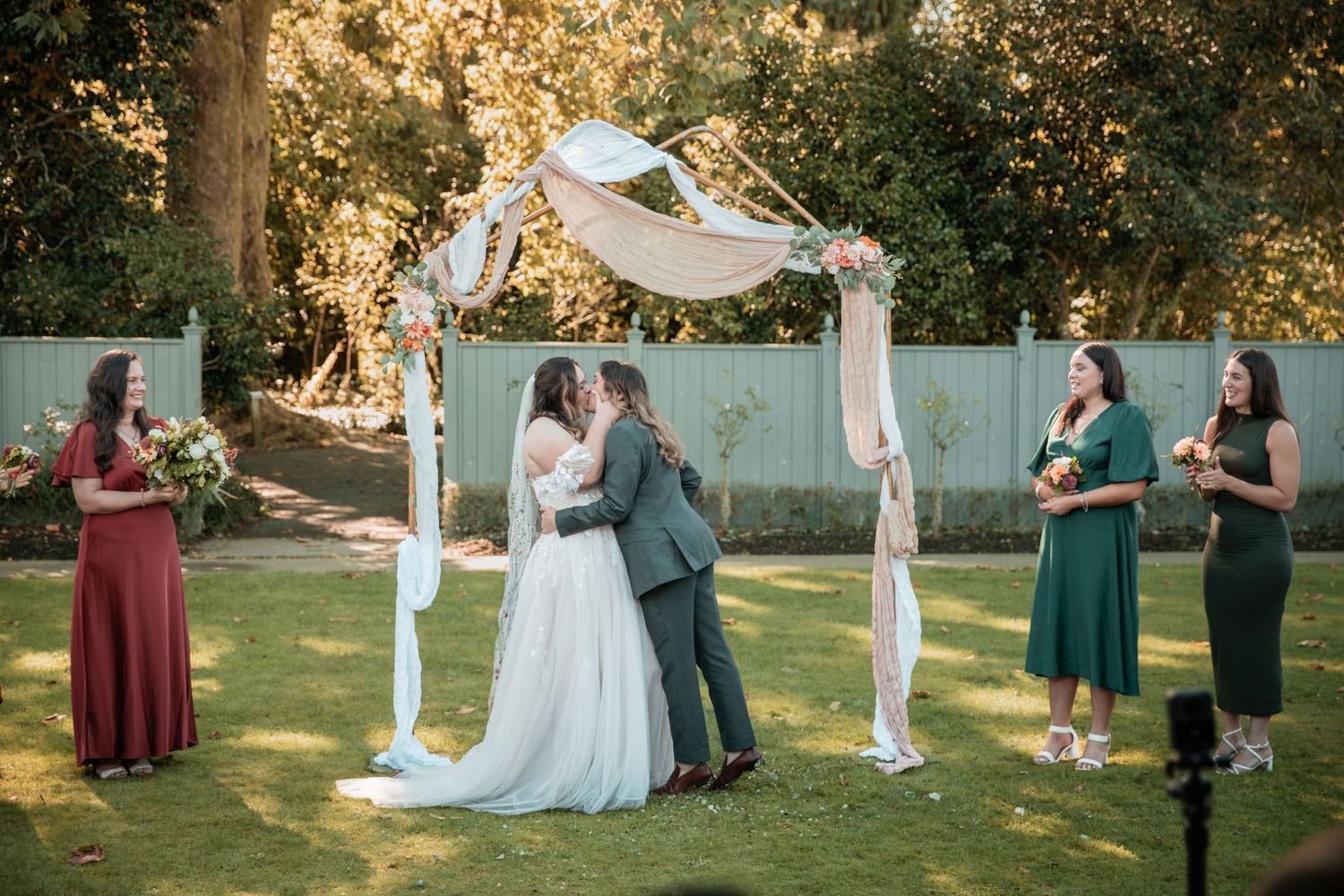 Couple shares first kiss under floral arch at outdoor wedding ceremony with bridesmaids in burgundy and green gowns.