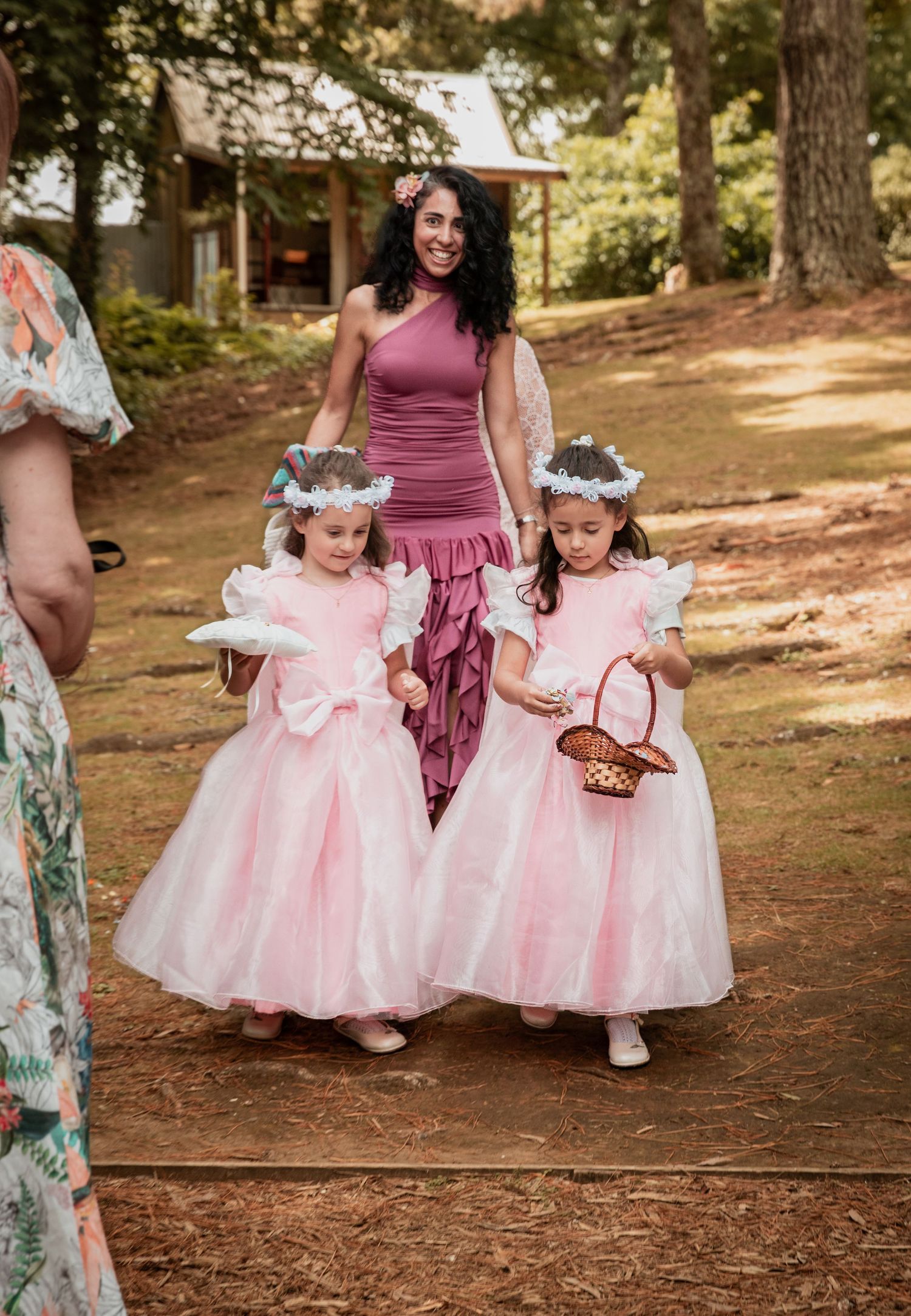 Two flower girls in pink dresses with floral crowns walk woodland path at outdoor wedding.