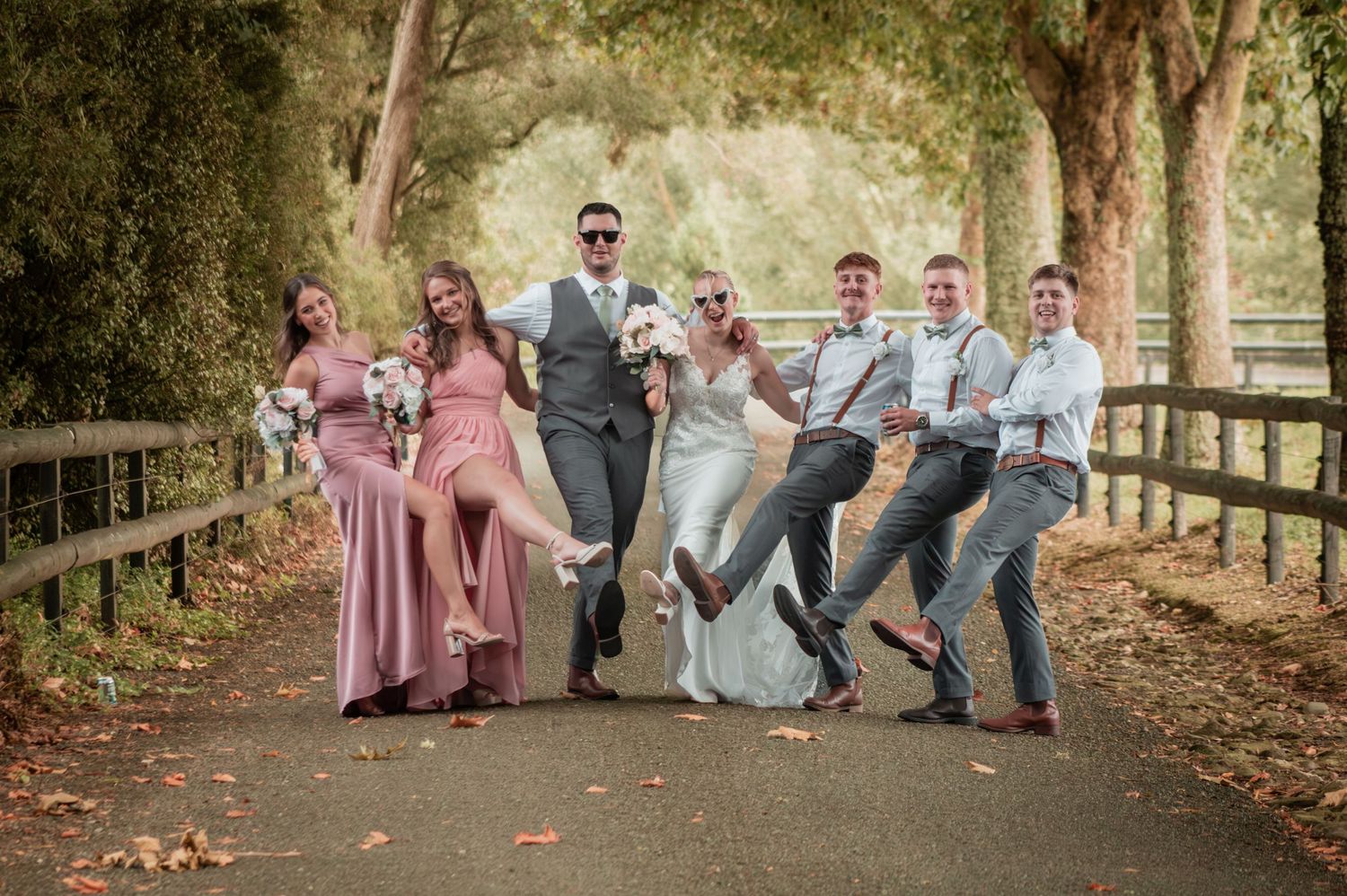 Wedding party laughs and poses playfully on tree-lined path in autumn setting.