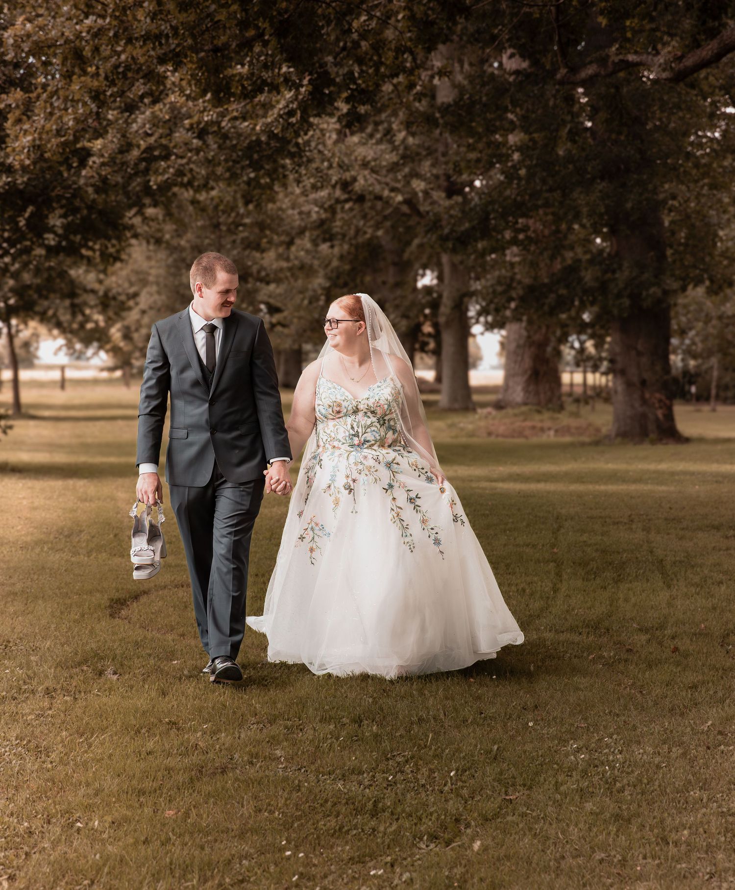 Wedding couple walking hand in hand across a lush green park with large trees in background.