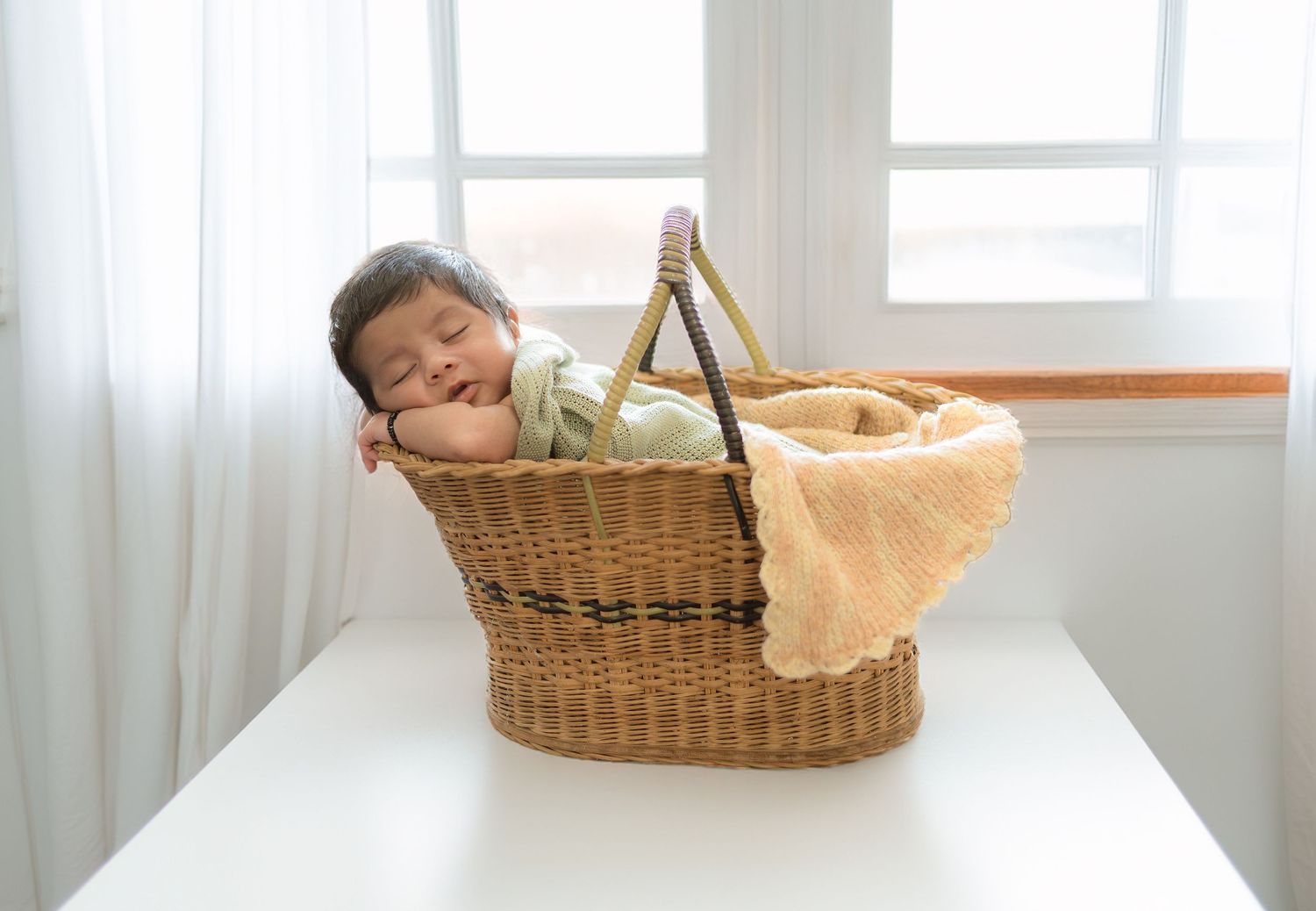 Sleeping newborn baby nestled in a wicker basket with a soft yellow blanket by a window.