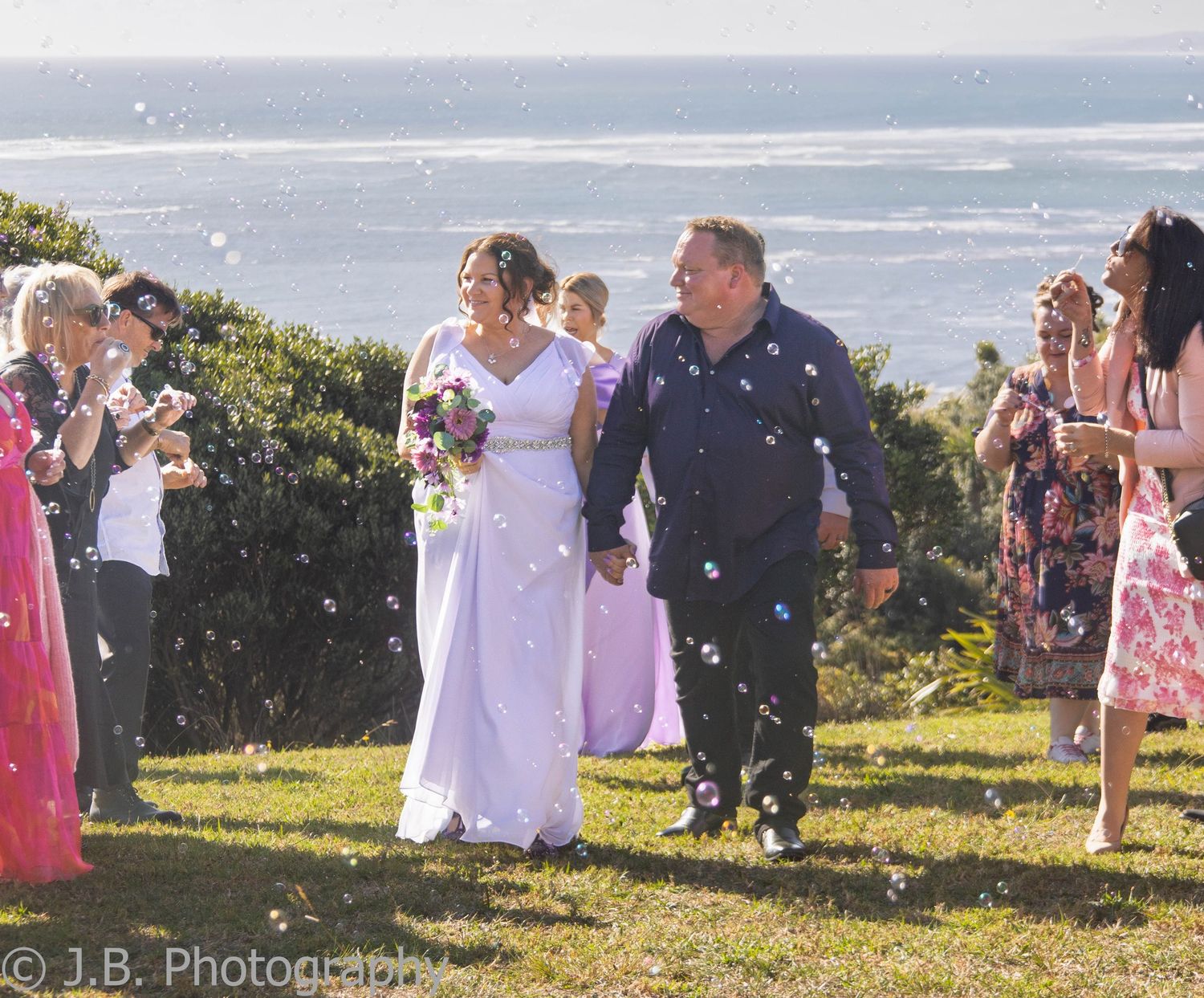 Bride and groom walking through bubbles with guests at an outdoor coastal wedding ceremony.