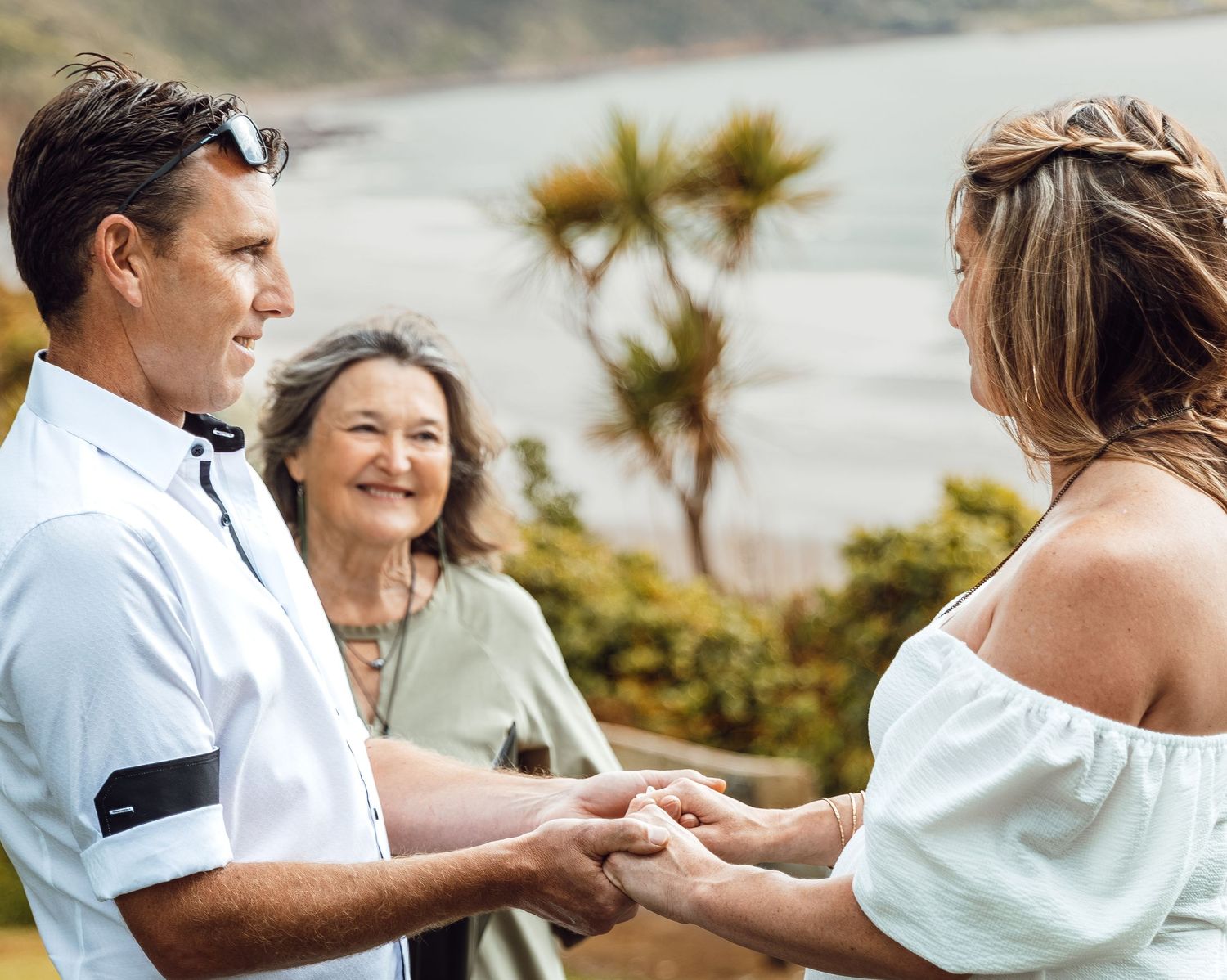 Couple exchanges vows holding hands with officiant and coastal scenery in background.