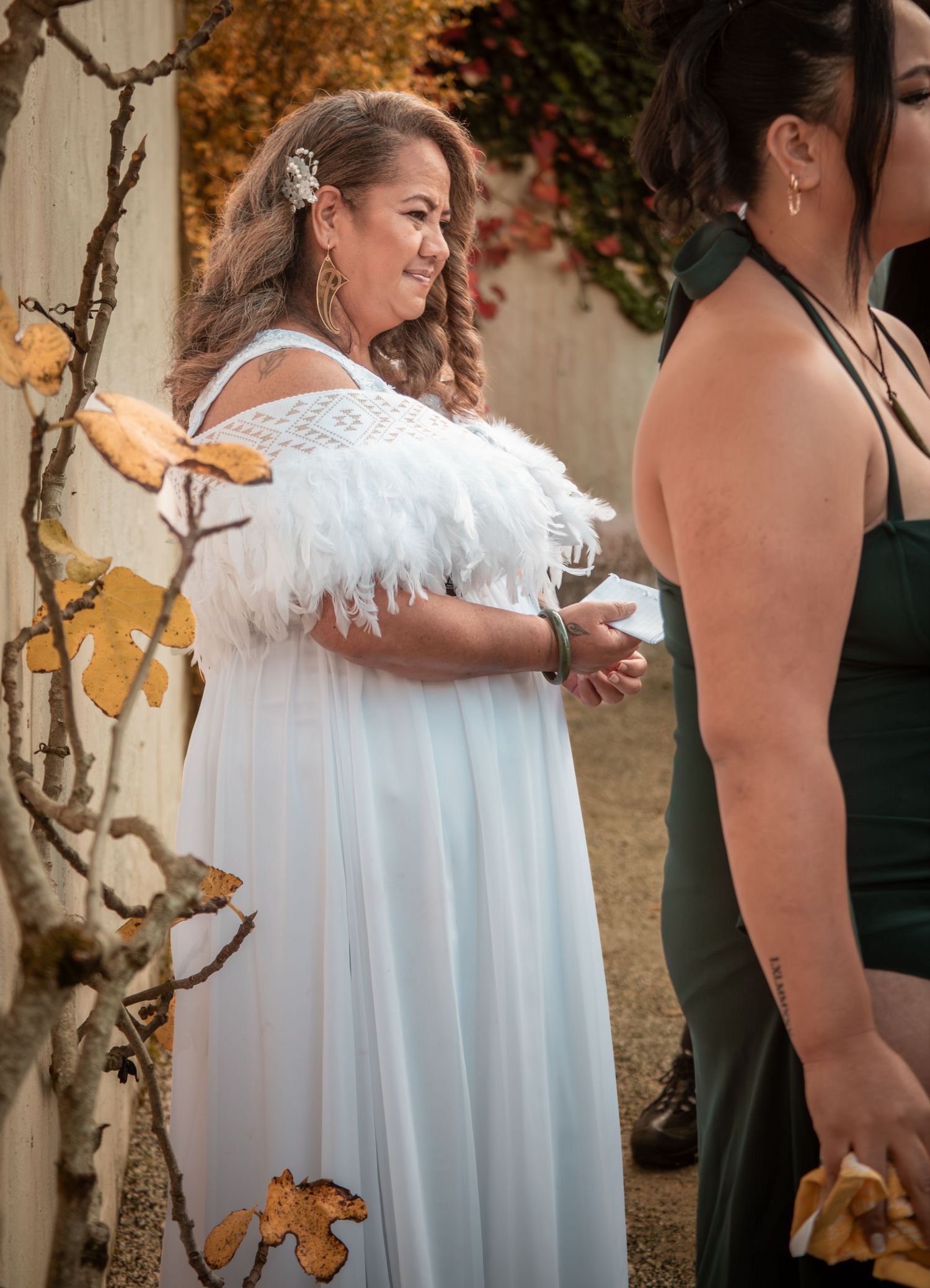 Woman in white feathered off-shoulder gown smiling at outdoor wedding ceremony with autumn foliage backdrop.