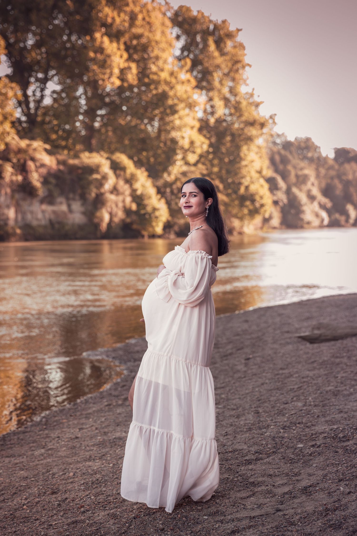 Pregnant woman in a white off-shoulder dress posing by a river surrounded by autumn trees.