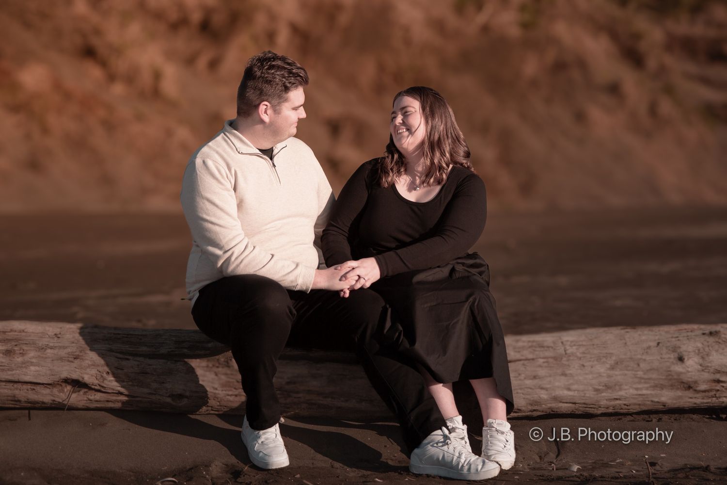 Couple sitting on driftwood holding hands and smiling at each other on a scenic beach.
