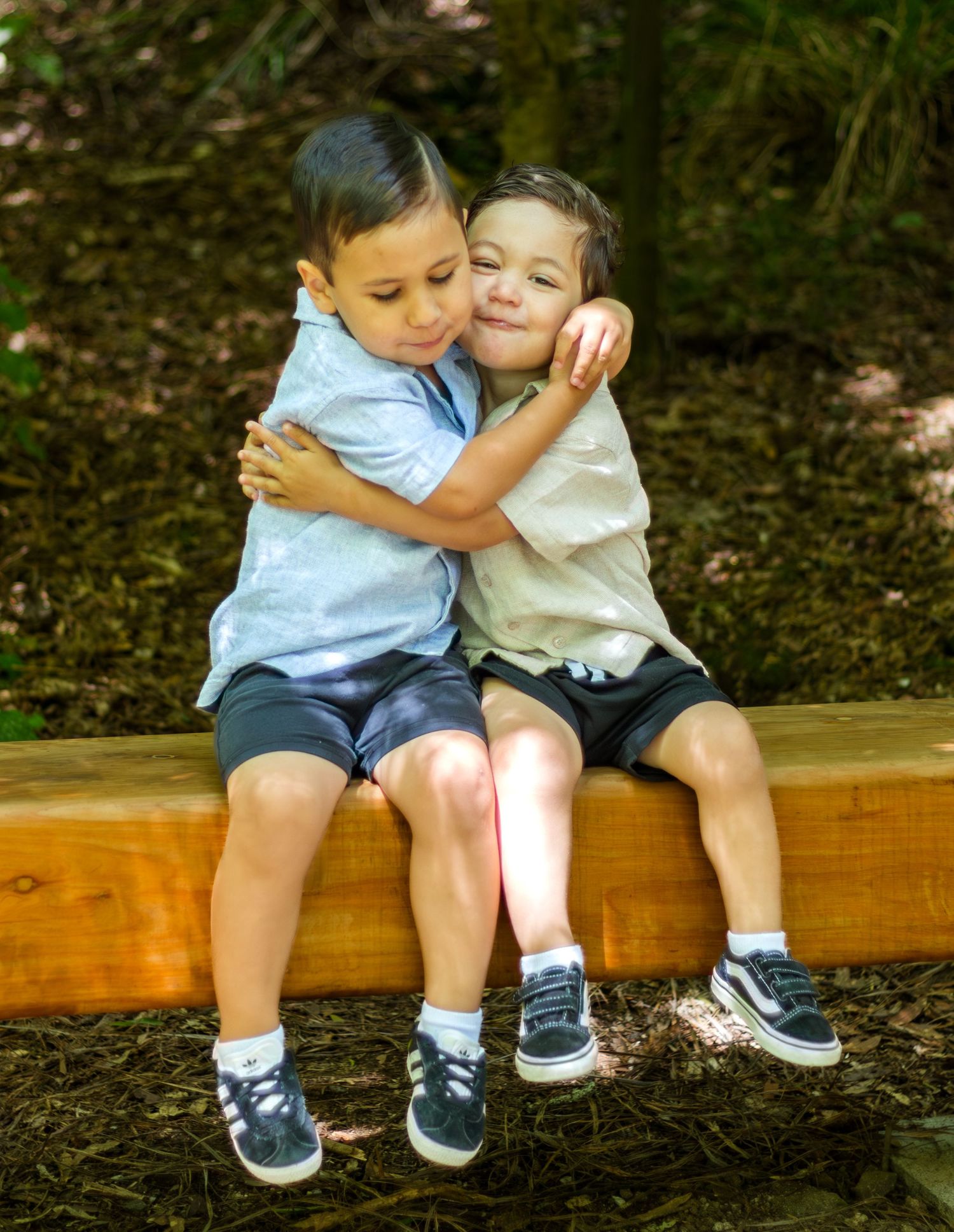 Two young boys share a sweet hug while sitting on a wooden bench outdoors.
