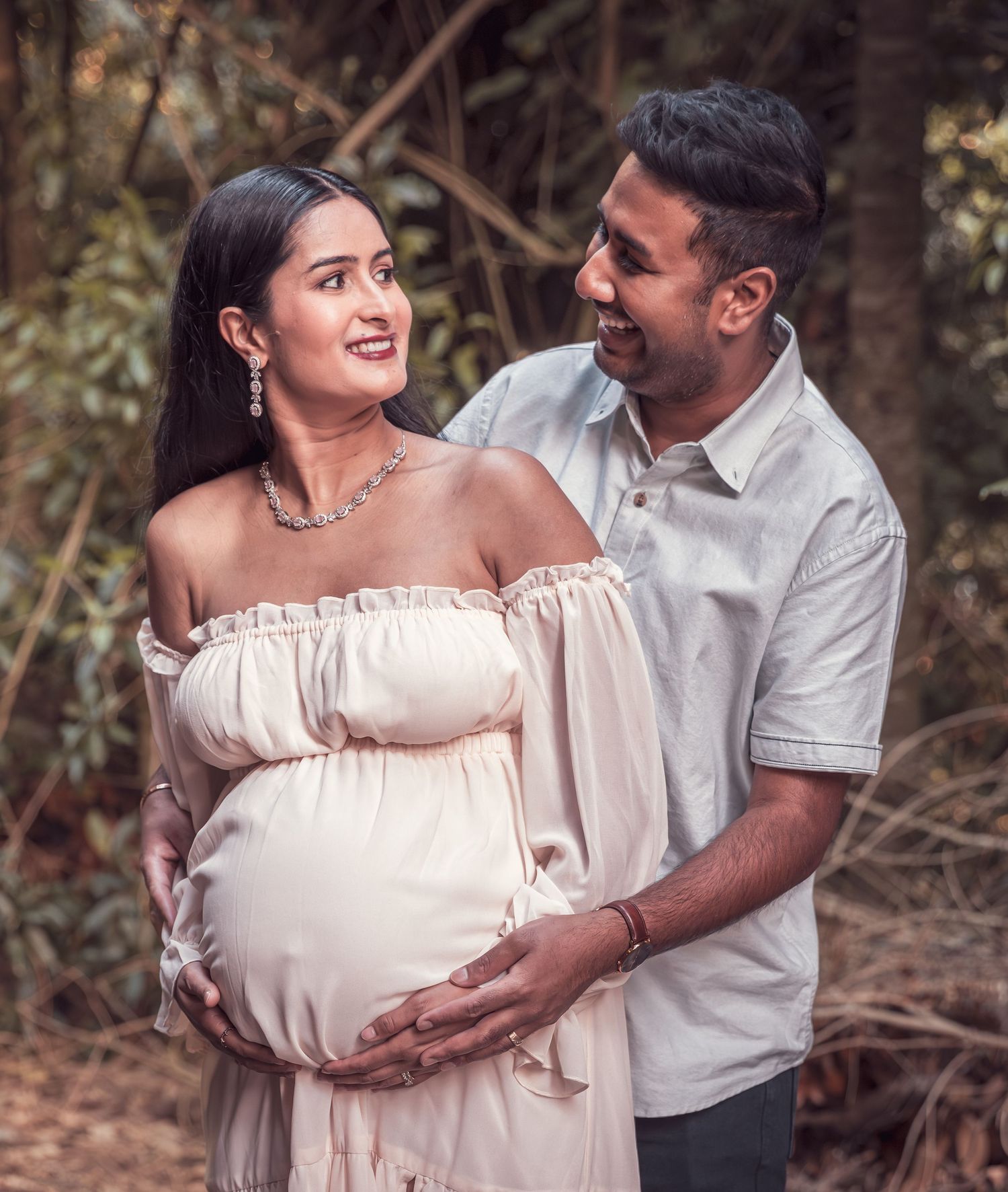 Happy couple posing for maternity photoshoot in forest, man tenderly holding pregnant womans baby bump.