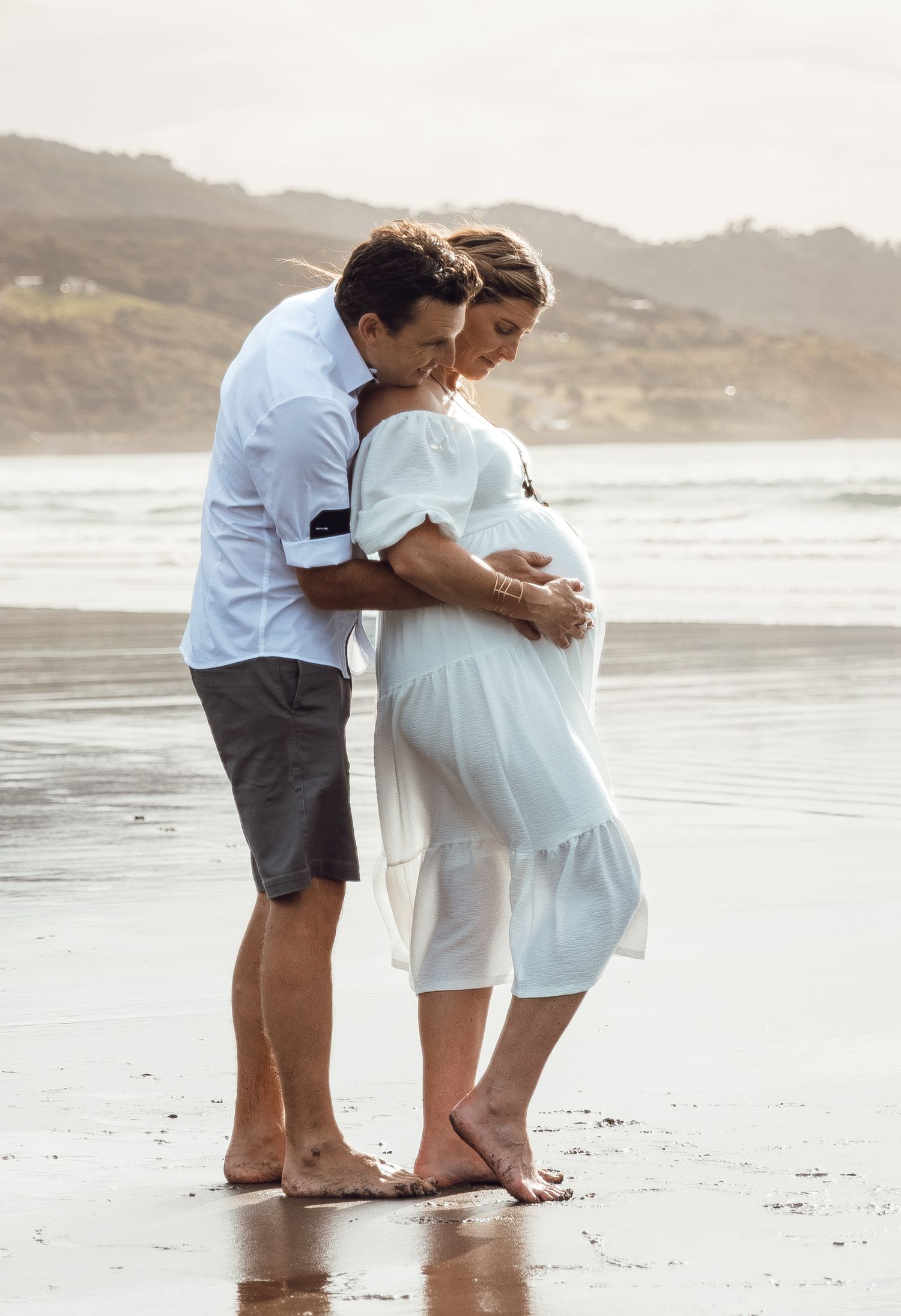Barefoot couple sharing tender moment on beach, man embracing pregnant woman in white dress by ocean.