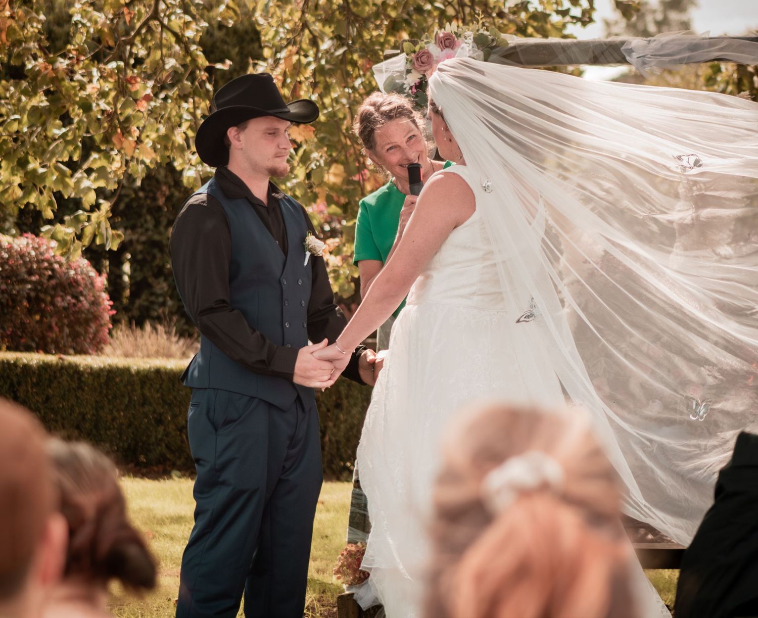 Groom in black hat holds brides hand as her veil blows in wind during garden ceremony.