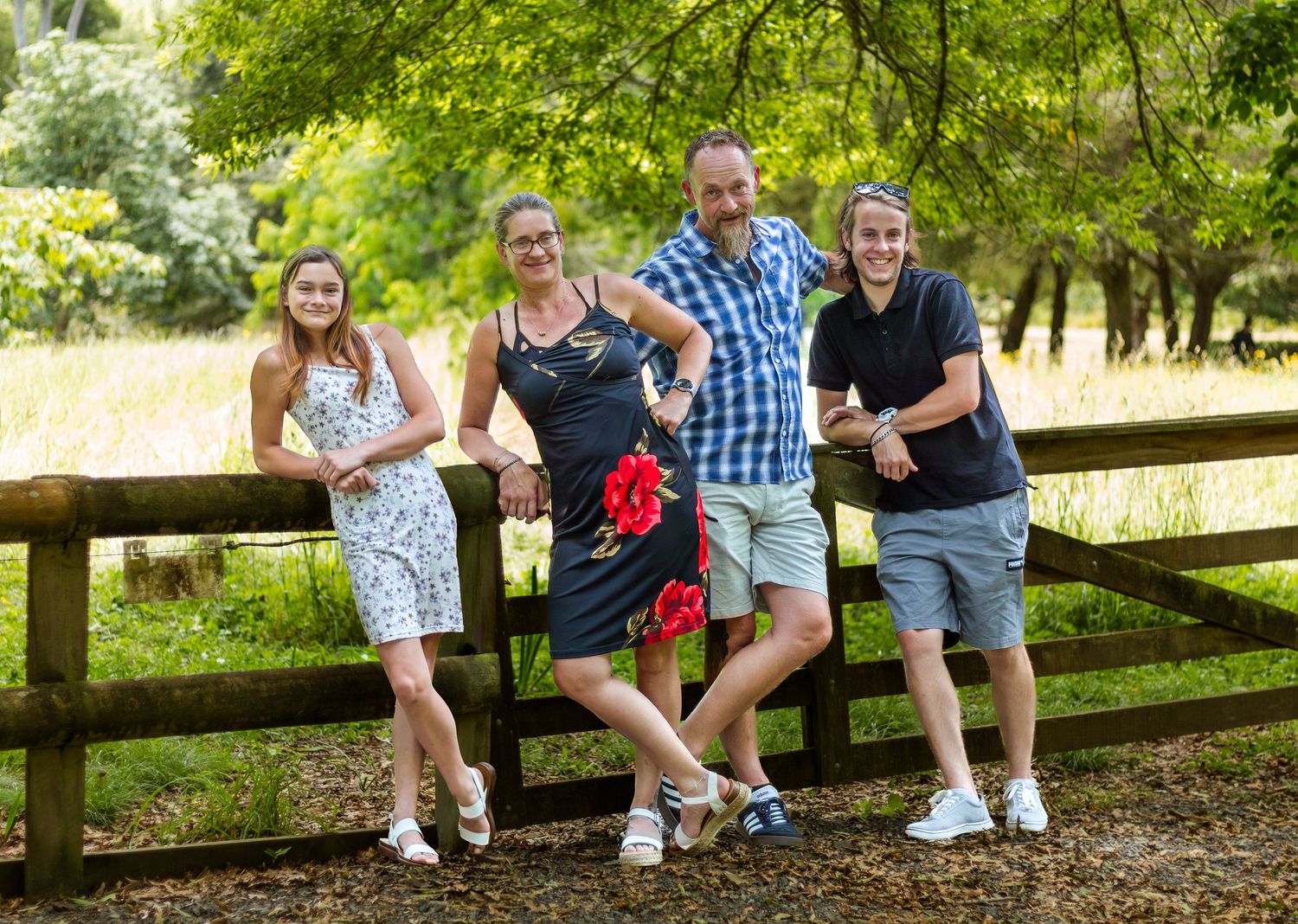 A happy family of four poses and laughs together leaning on a wooden fence outdoors.