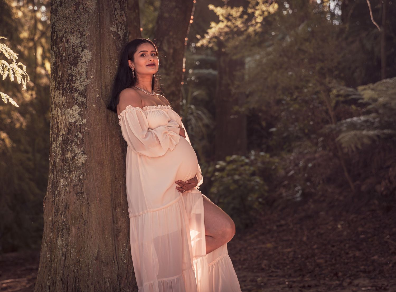 Elegant pregnant woman in flowing white off-shoulder gown leaning against tree in enchanted forest setting.