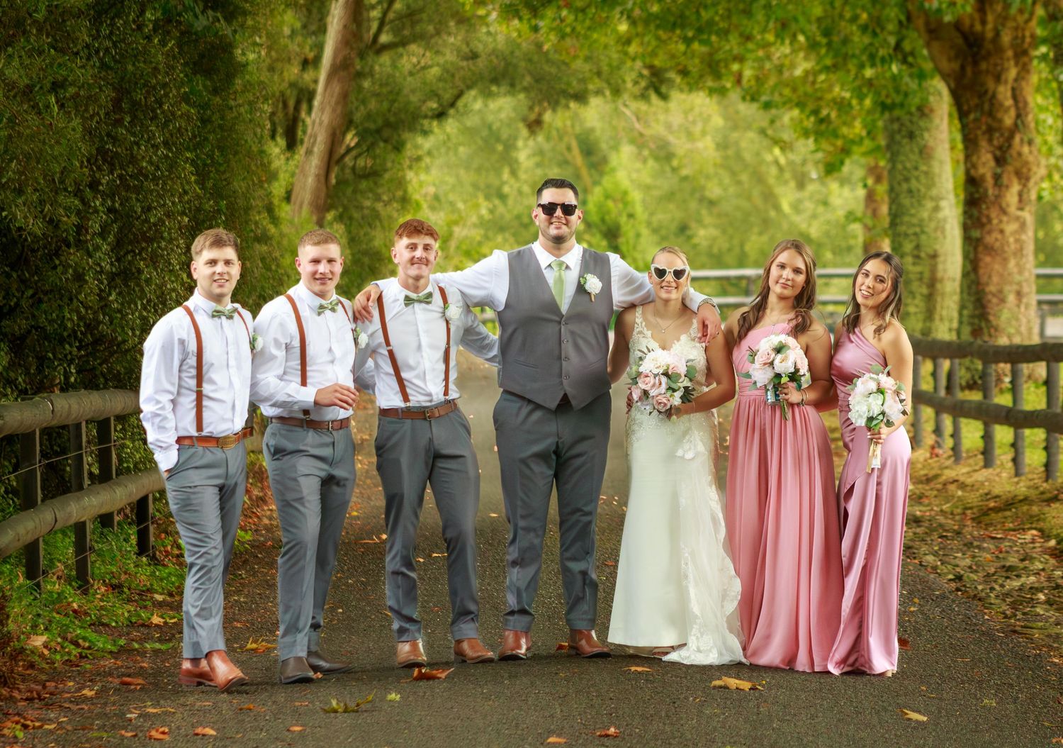 Wedding party in grey suits and pink bridesmaid gowns posing on a tree-lined country lane.