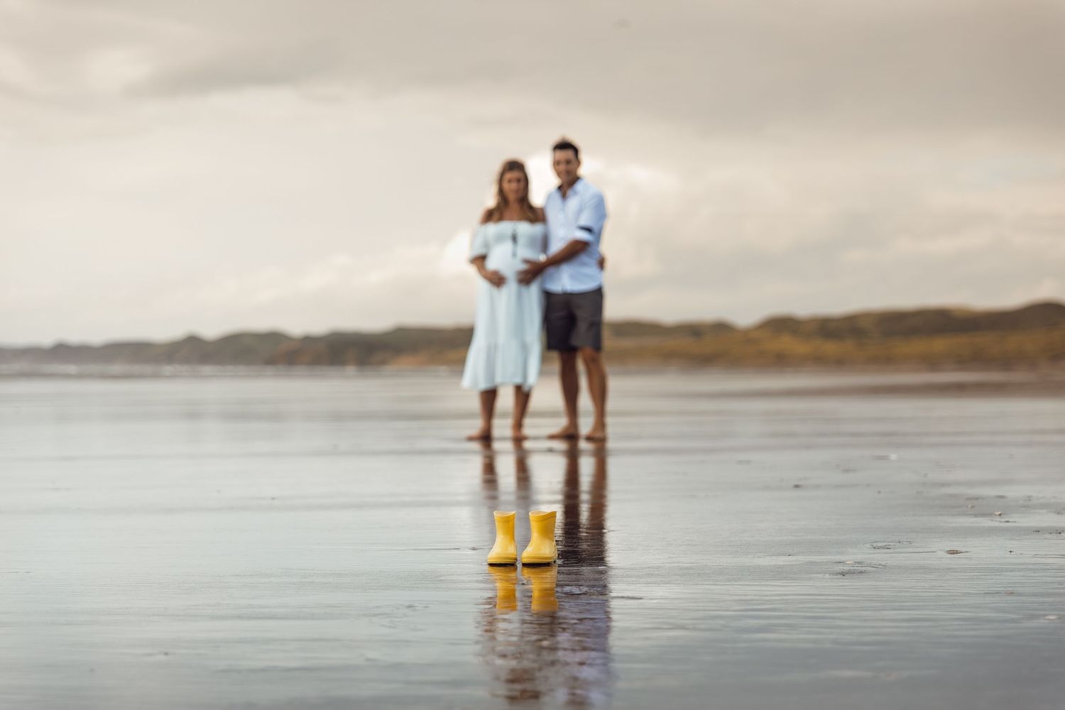 Expectant couple standing together on a reflective beach with yellow rain boots placed in front of them.