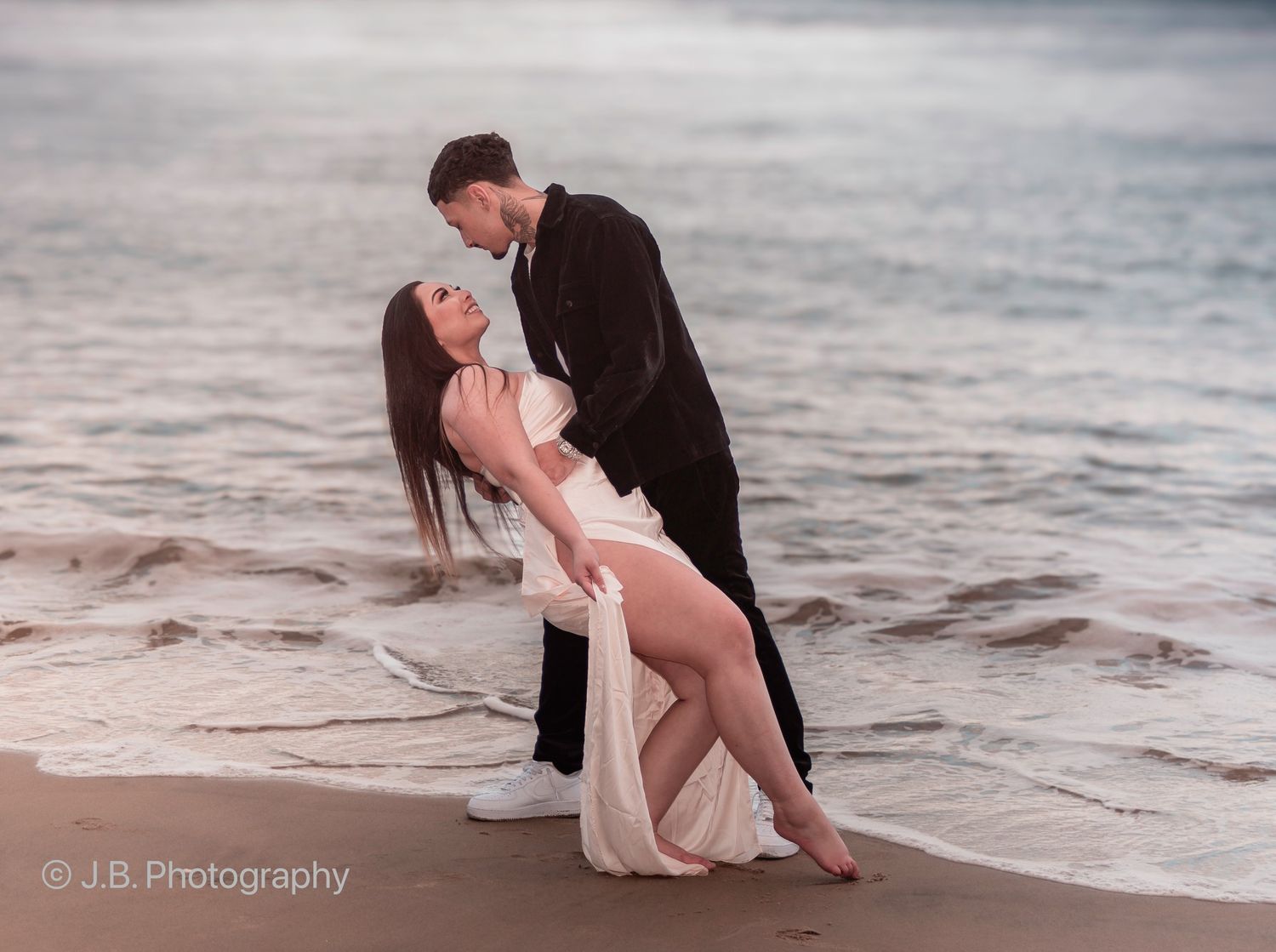 Romantic couple dancing on a beach at sunset, man dipping woman in elegant white dress by the ocean waves.