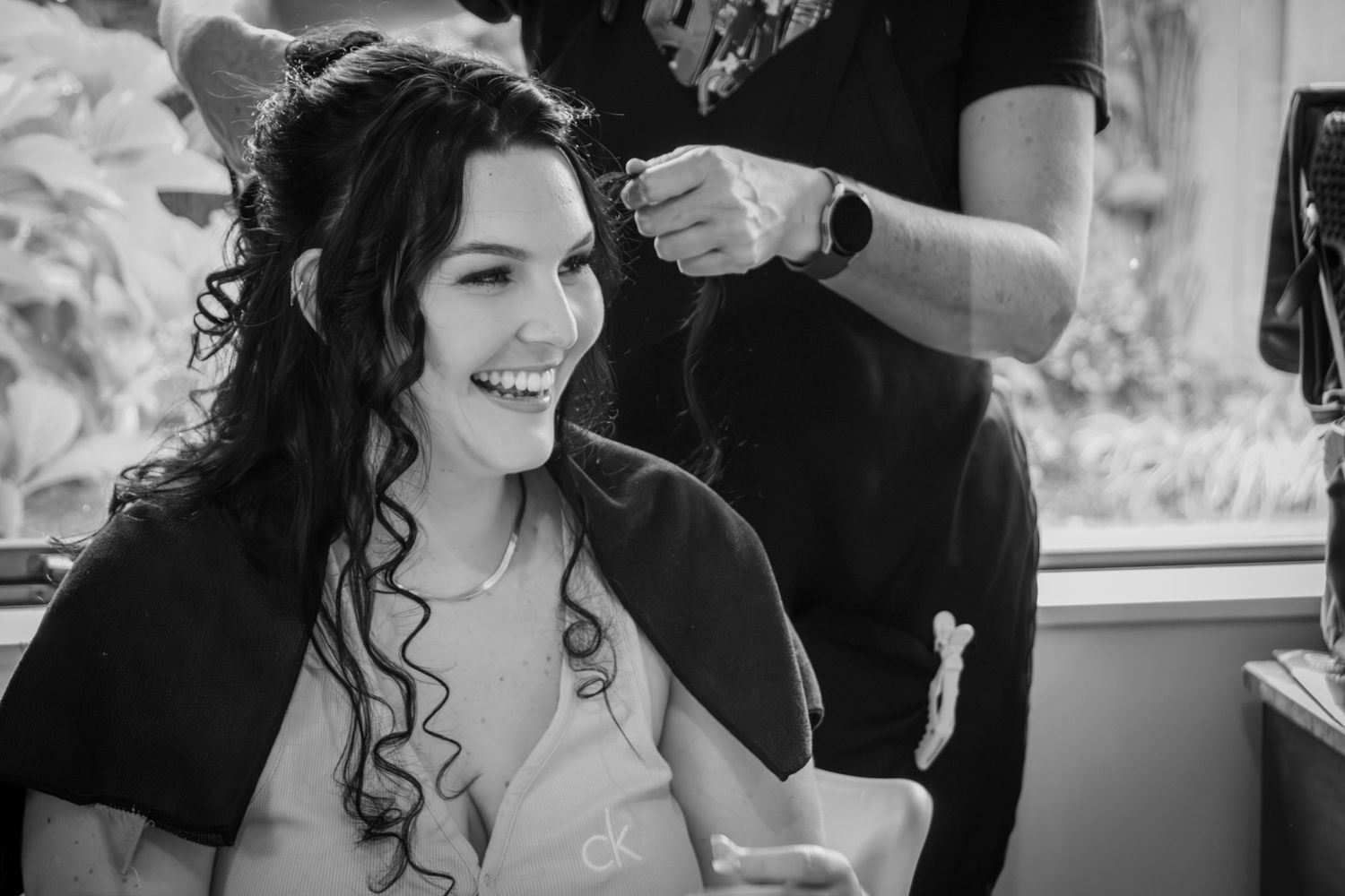 Bride smiling joyfully in black and white photo while getting her hair styled before the wedding ceremony.
