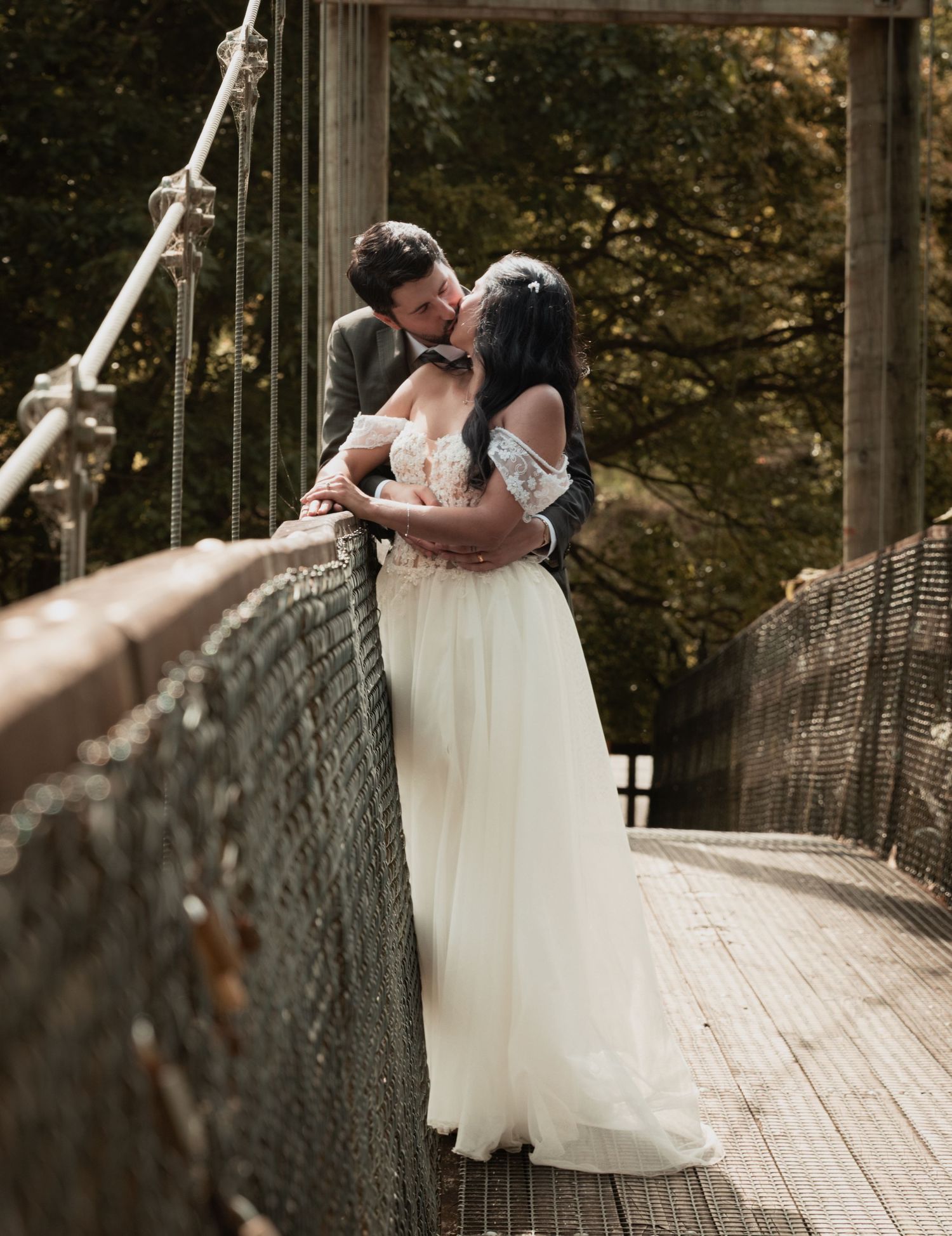 Bride and groom sharing a romantic kiss on a rustic suspension bridge surrounded by autumn trees.