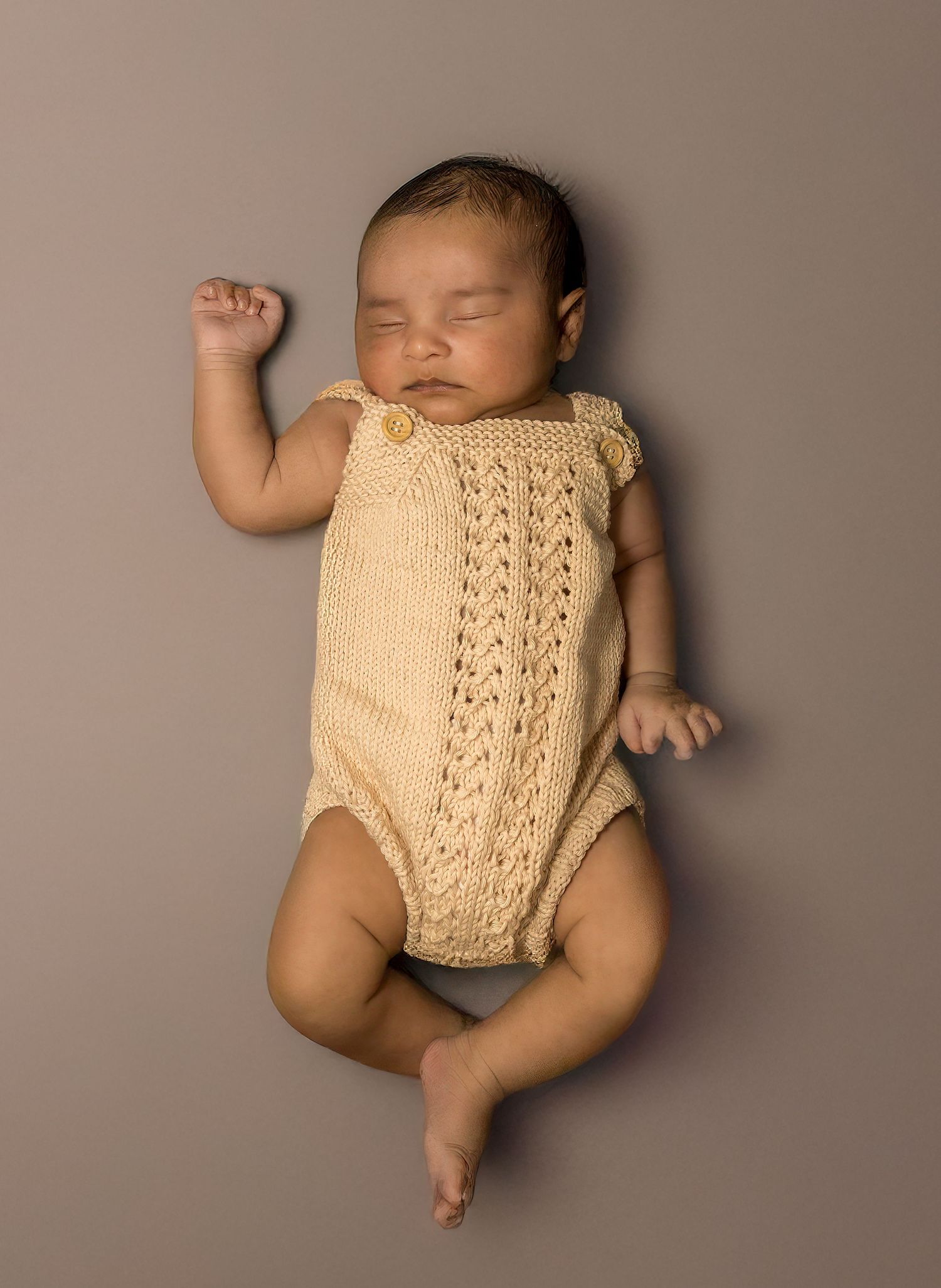 Sleeping newborn baby wearing a cream knitted romper, lying peacefully on a taupe neutral background.