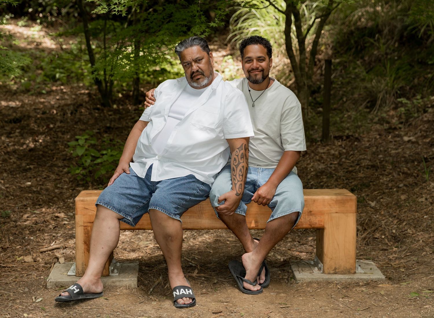 Two men smile warmly sitting together on a wooden bench in a lush outdoor setting.
