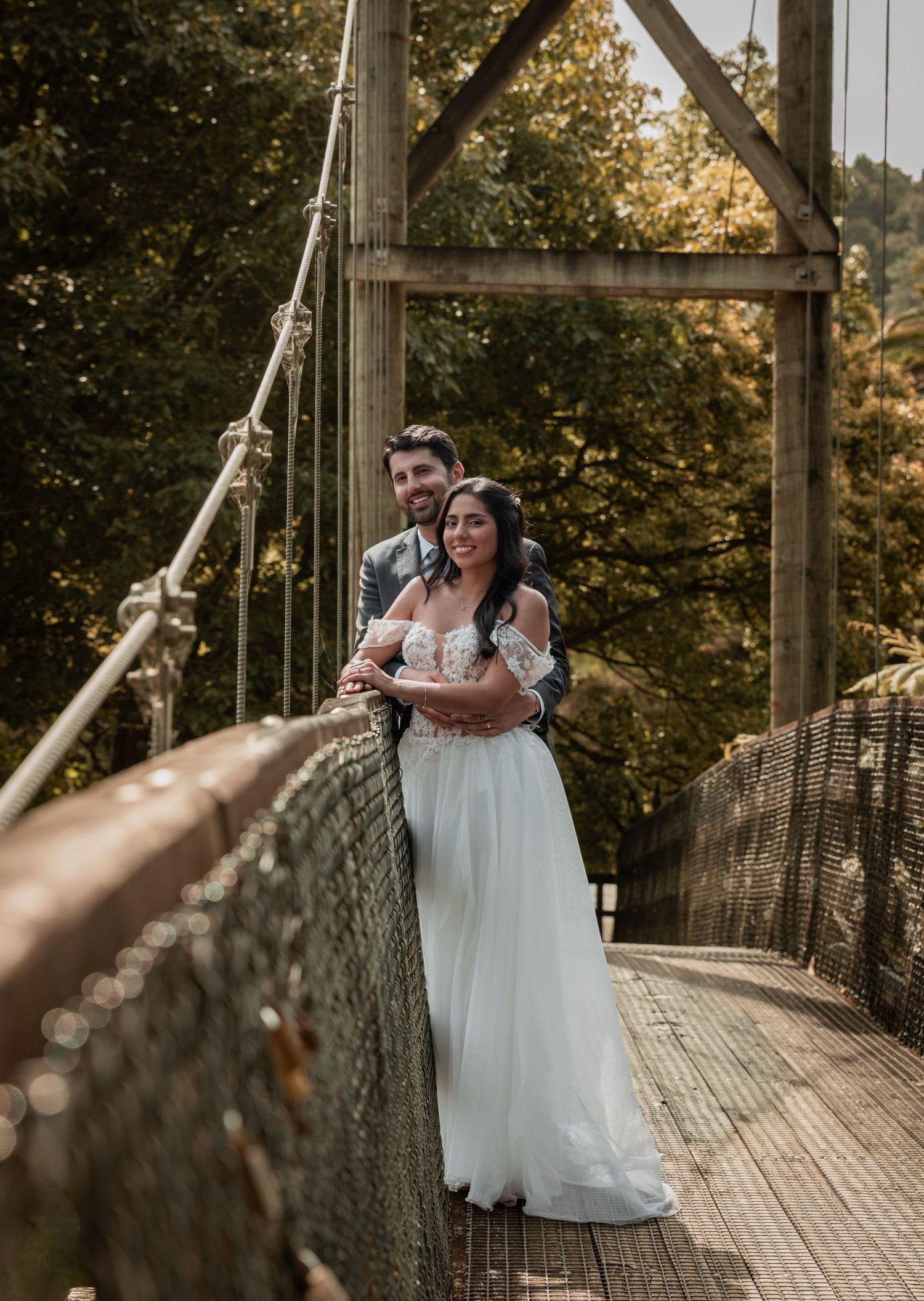 Bride and groom embracing on rustic suspension bridge surrounded by autumn foliage in wedding portrait.