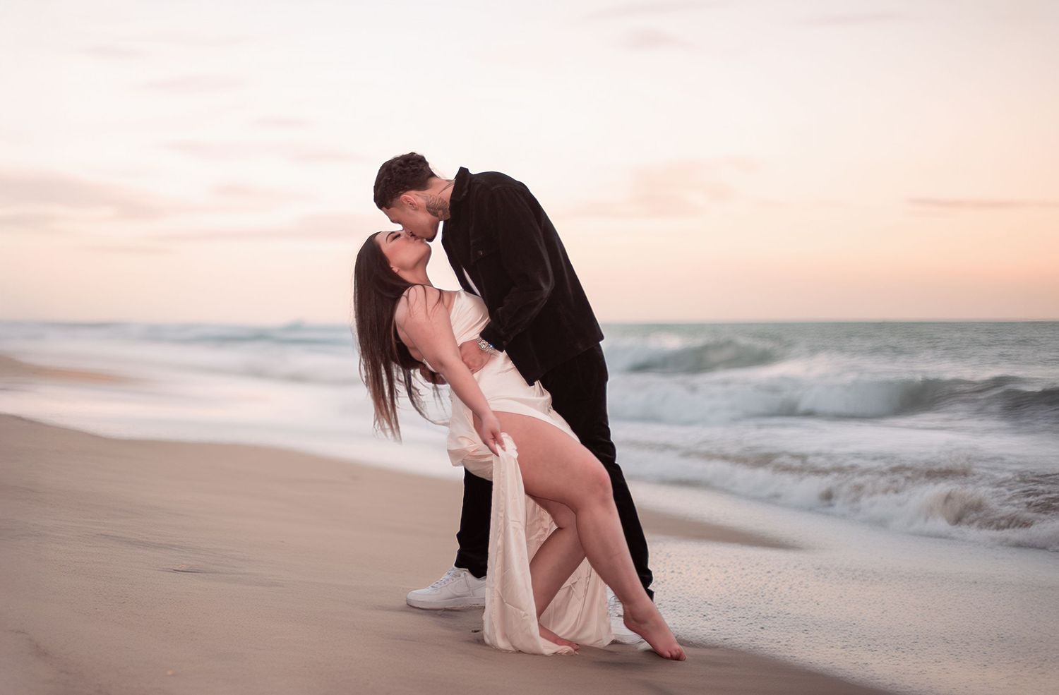 Couple sharing romantic dip kiss on sandy beach at sunset with ocean waves in background.