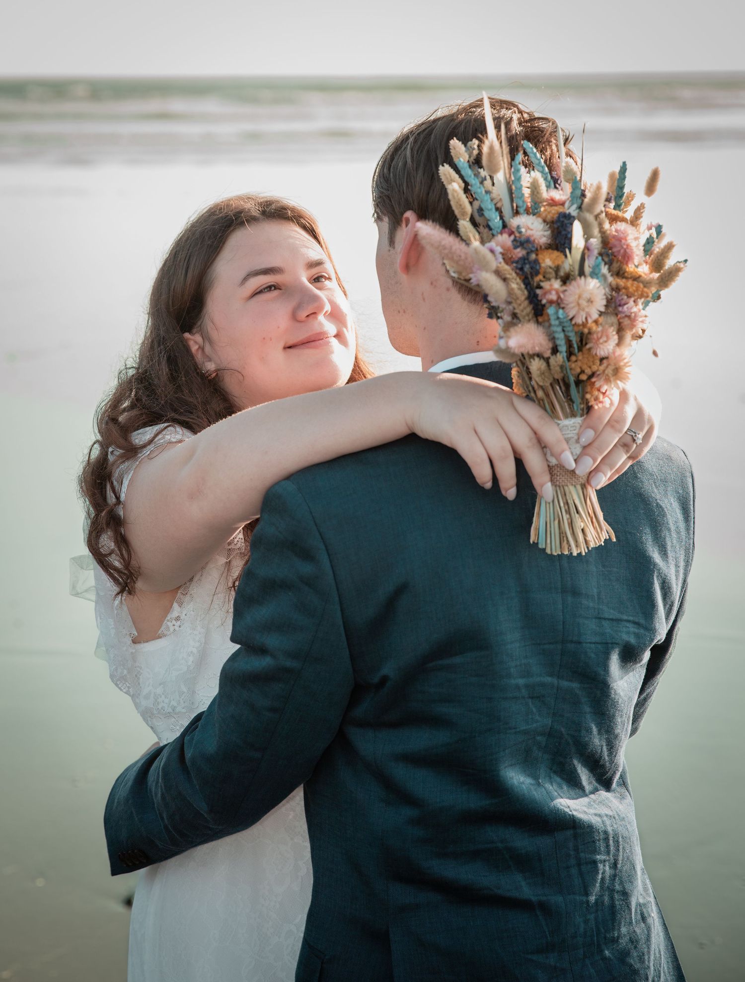 Bride smiles holding colorful dried flower bouquet while embracing groom on beach at sunset.
