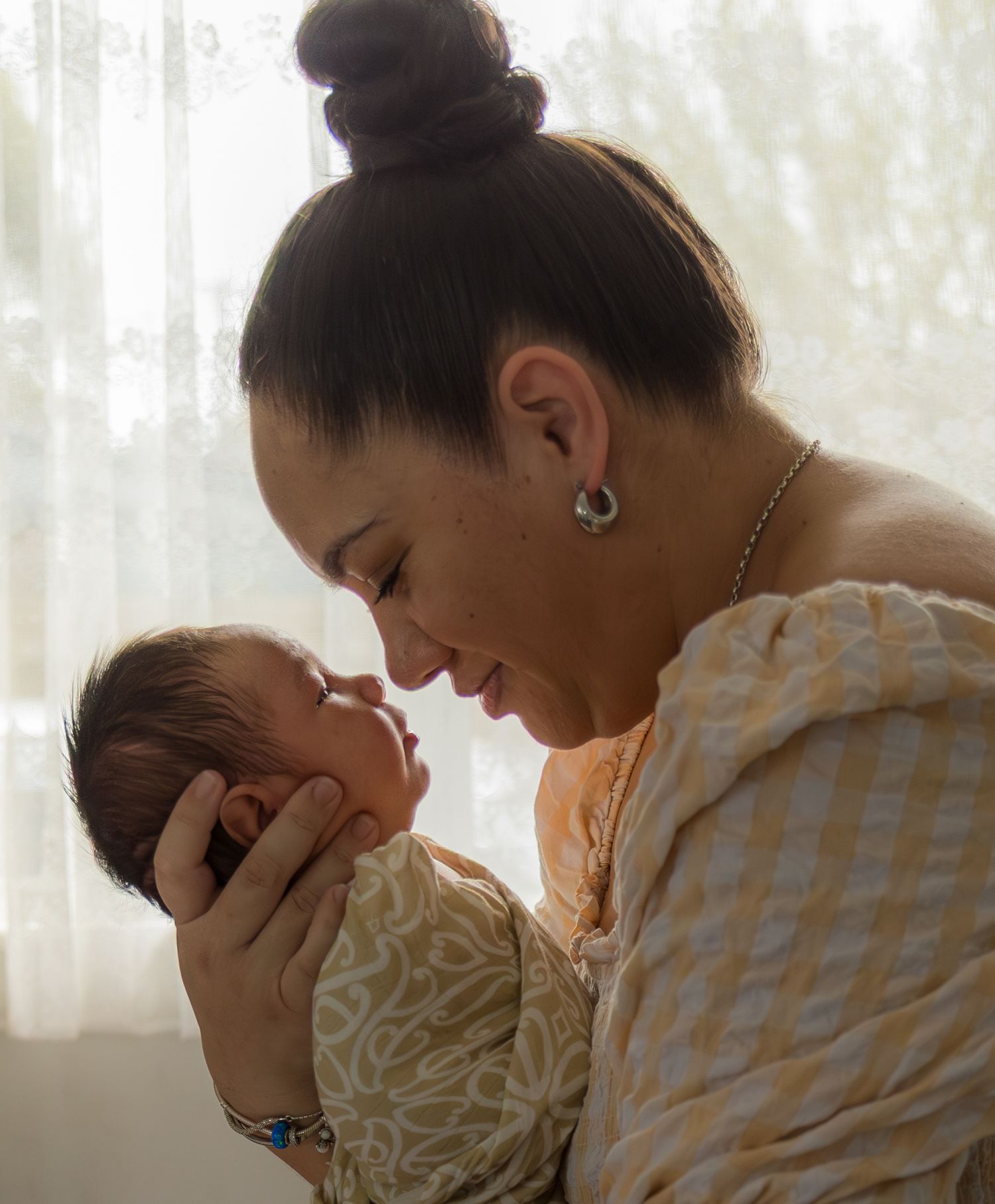 Mother with hair in bun tenderly holding newborn baby close to her face near a soft curtained window.