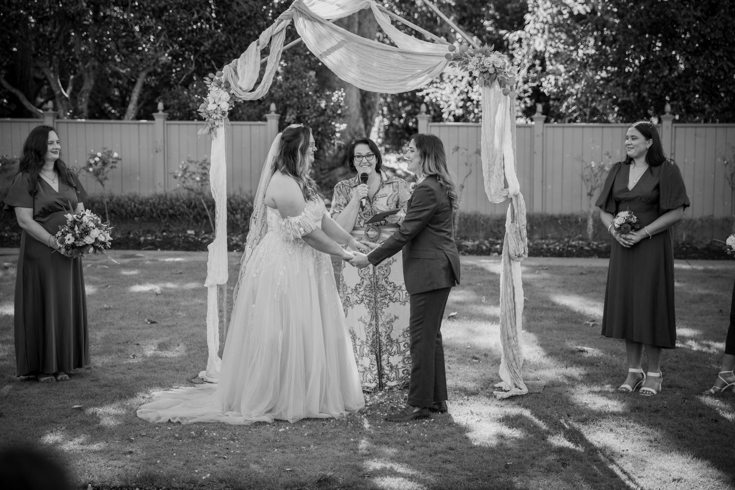 Bride and groom exchanging vows under floral arch with bridesmaids, black and white outdoor ceremony.