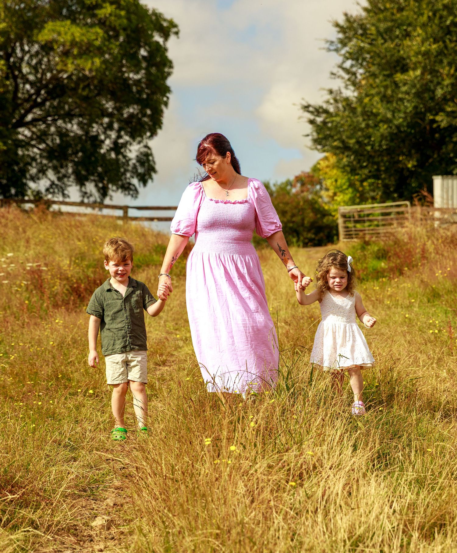 A mother in a pink dress walks through a sunny meadow holding hands with her two young children.