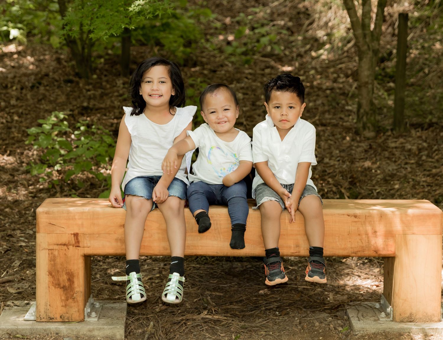 Three young siblings in white tops sit together smiling on a wooden bench outdoors.