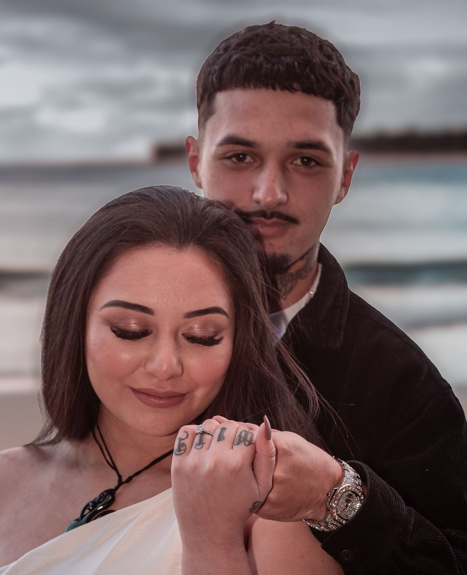 Couple embracing on beach at dusk with tattooed hands intertwined in romantic portrait.