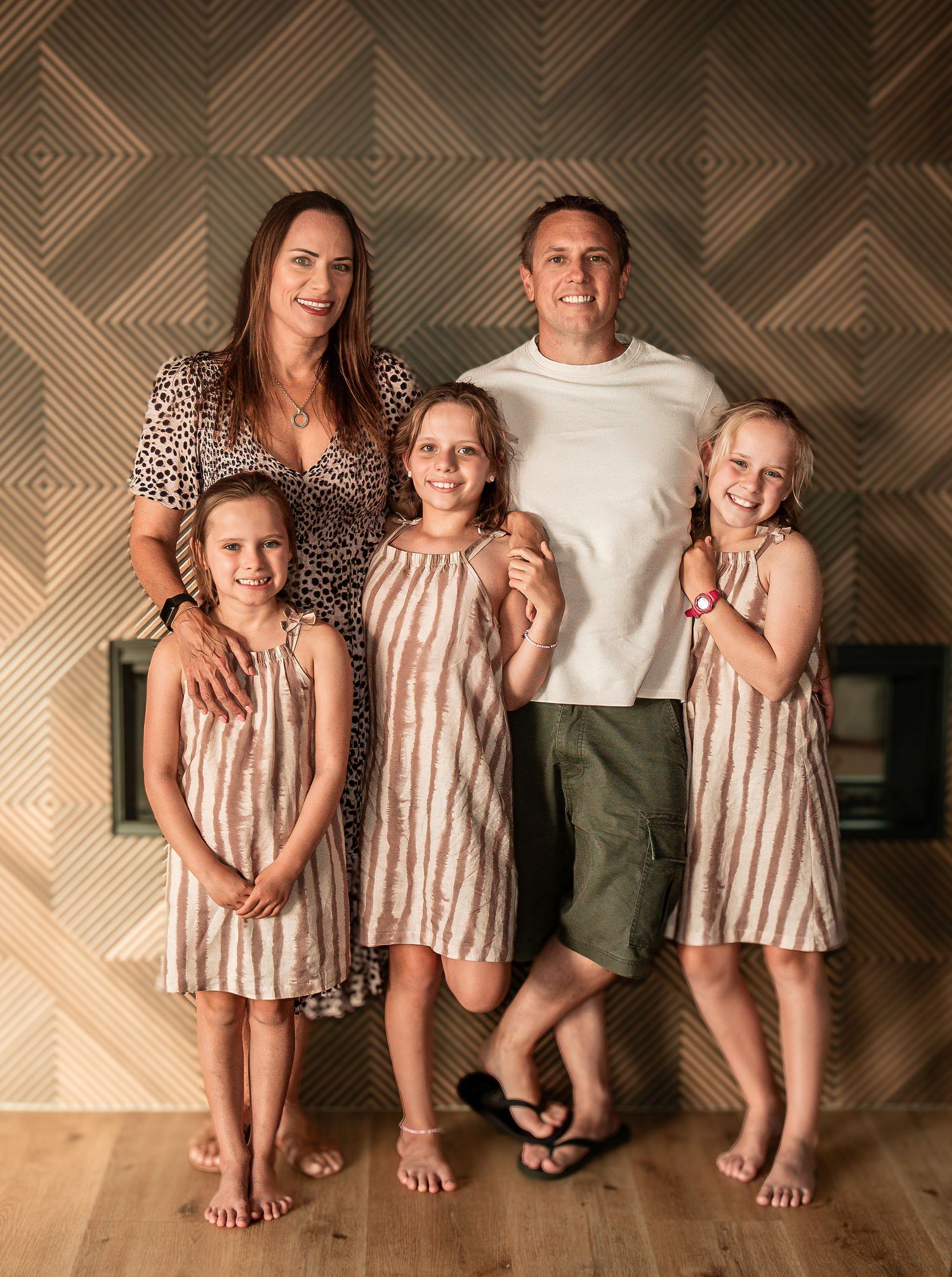 A family of five with three daughters in matching dresses poses indoors against a patterned wall.