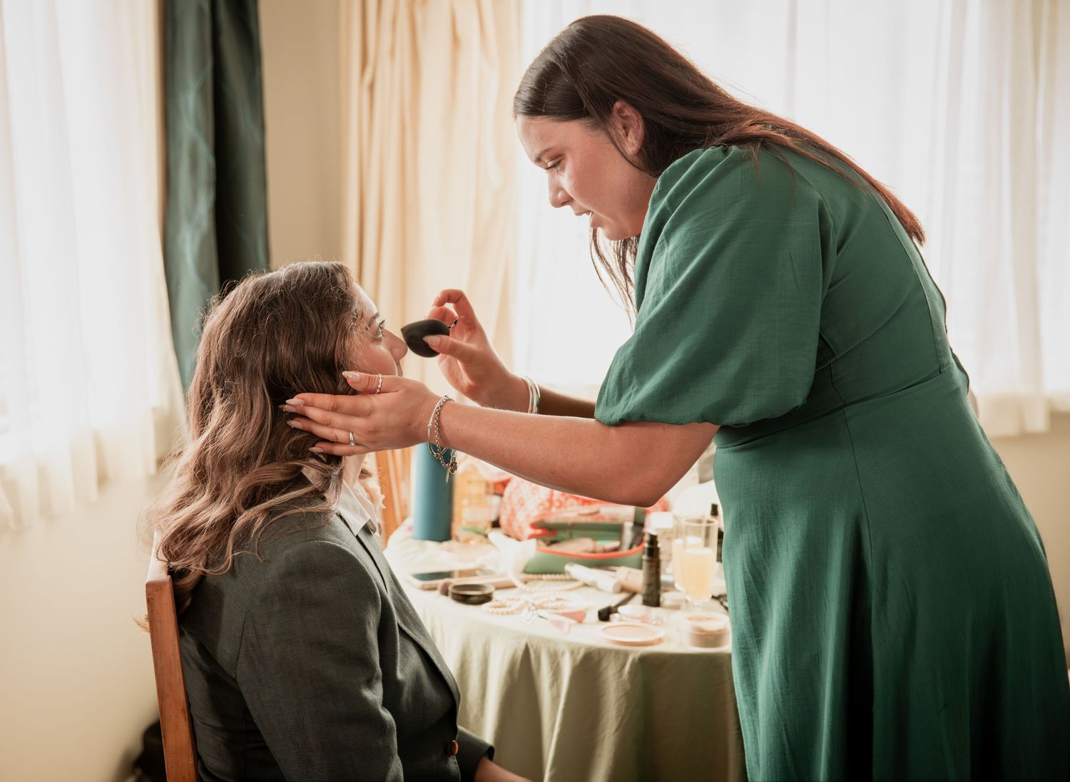 Makeup artist in green dress applies cosmetics to seated woman near window in bright room.