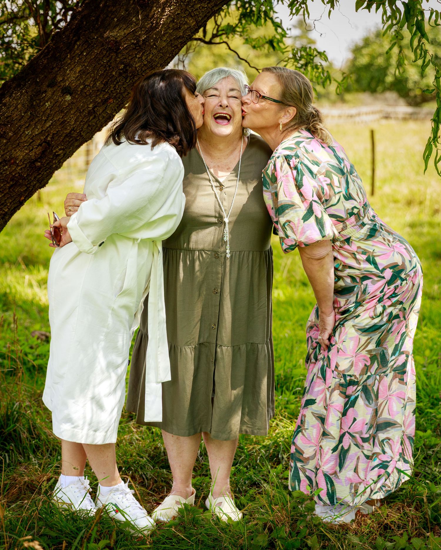 Three women sharing joyful moment with kisses under tree in outdoor family portrait.