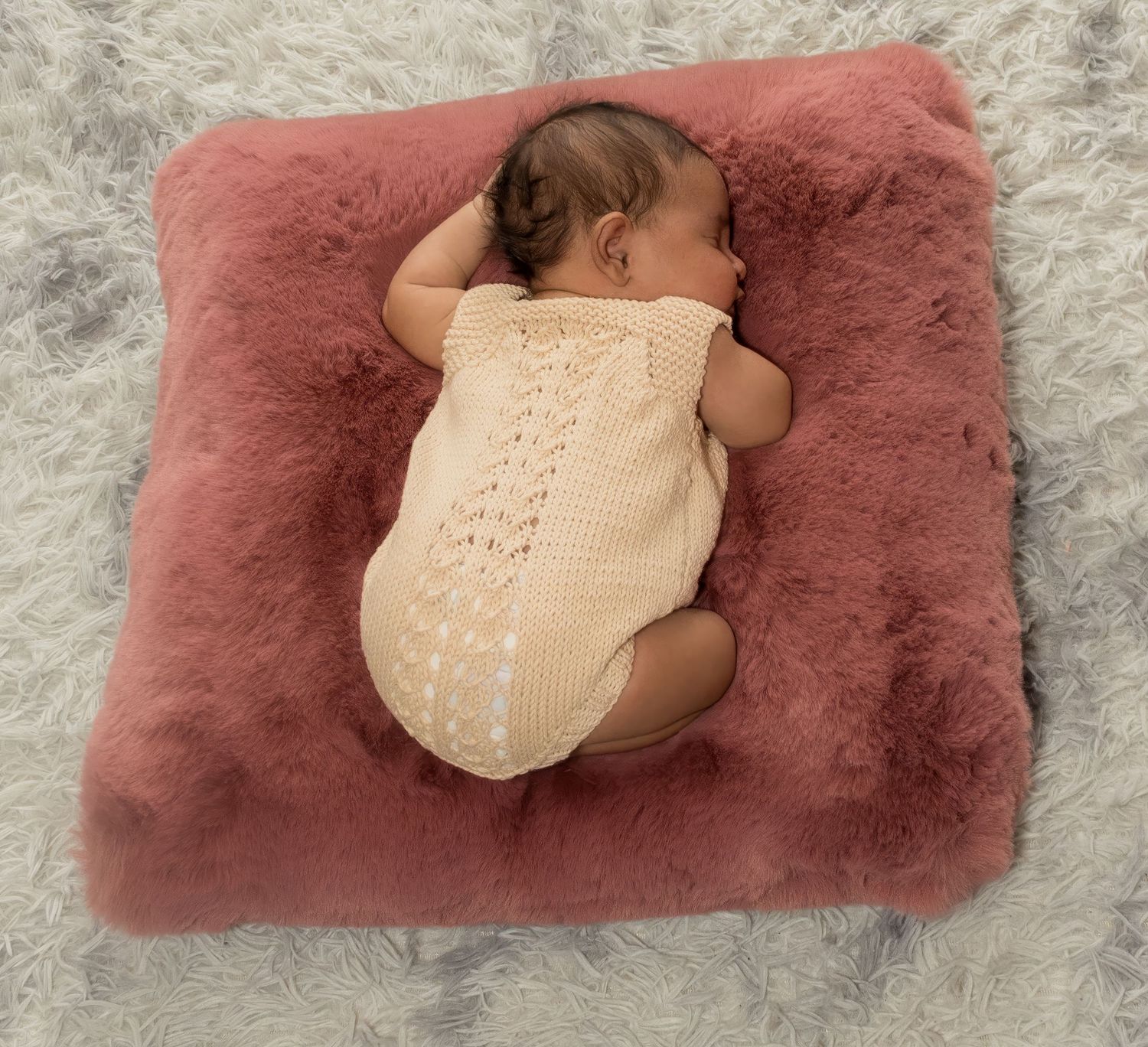 Newborn baby sleeping peacefully on a pink fluffy blanket wearing a cream knit romper.