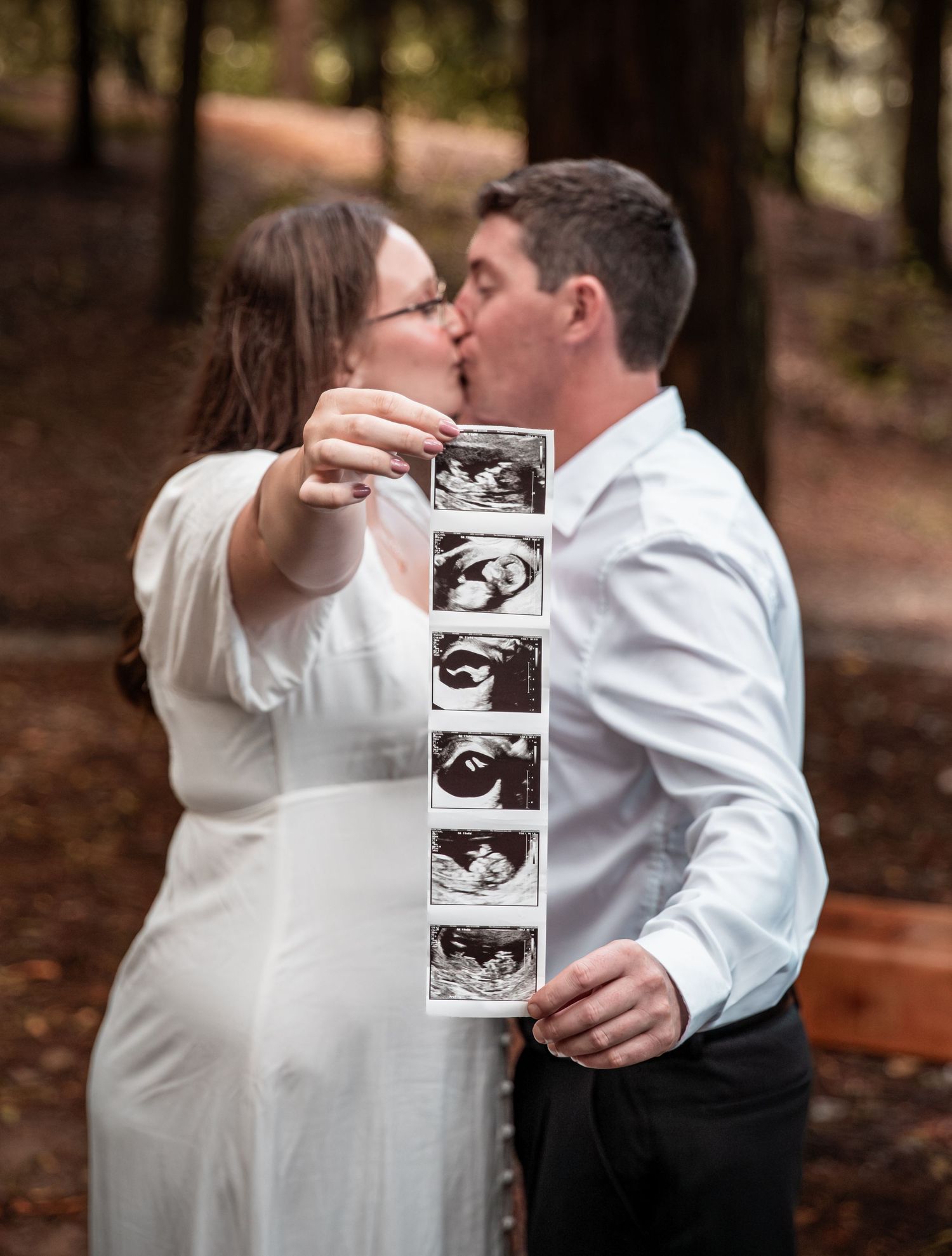 Excited couple kissing while holding ultrasound photo strip during outdoor pregnancy announcement photoshoot.